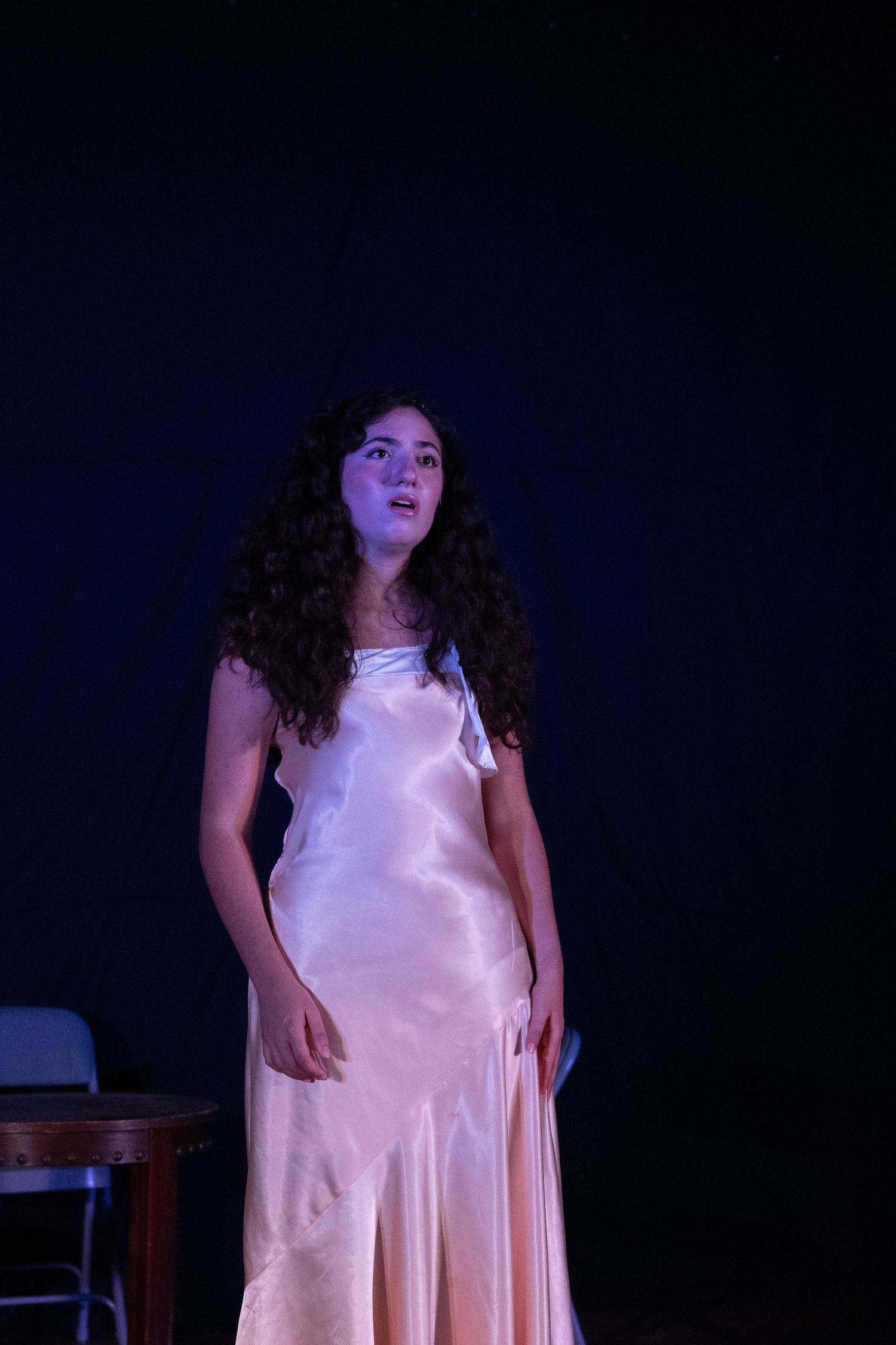 Young woman in a satin pink gown performing on a dark stage under blue and purple theatrical lighting.