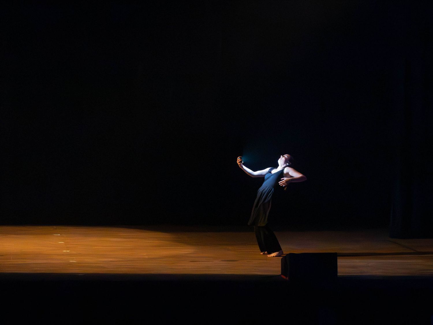 A lone dancer in a dark dress leans backward with outstretched arms on a dimly lit wooden stage, illuminated by a soft spotlight.