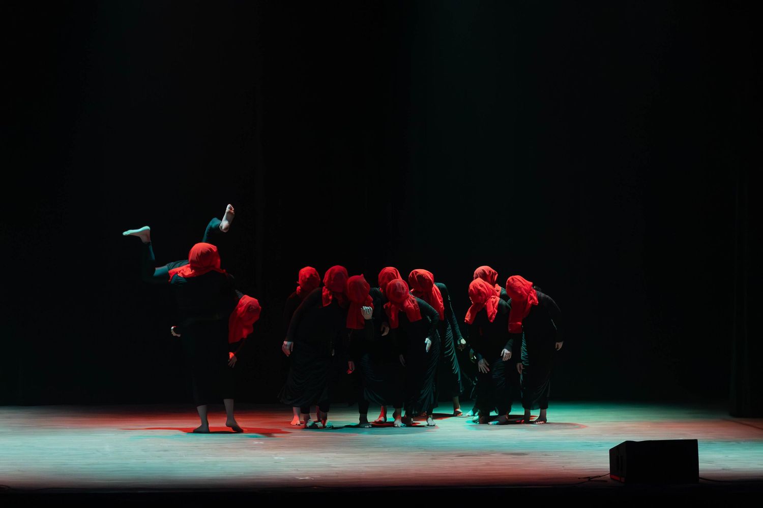 Performers in red hoods and black costumes huddle together on a dimly lit stage during a dramatic theatrical performance.