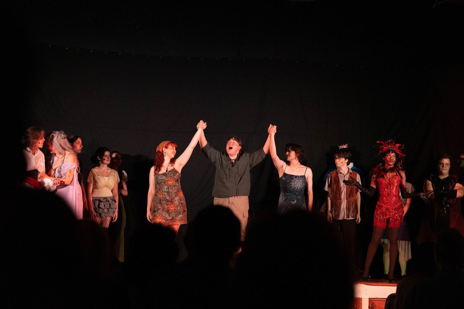 Theater performers take a bow on stage during a curtain call, with cast members holding hands under dramatic lighting.