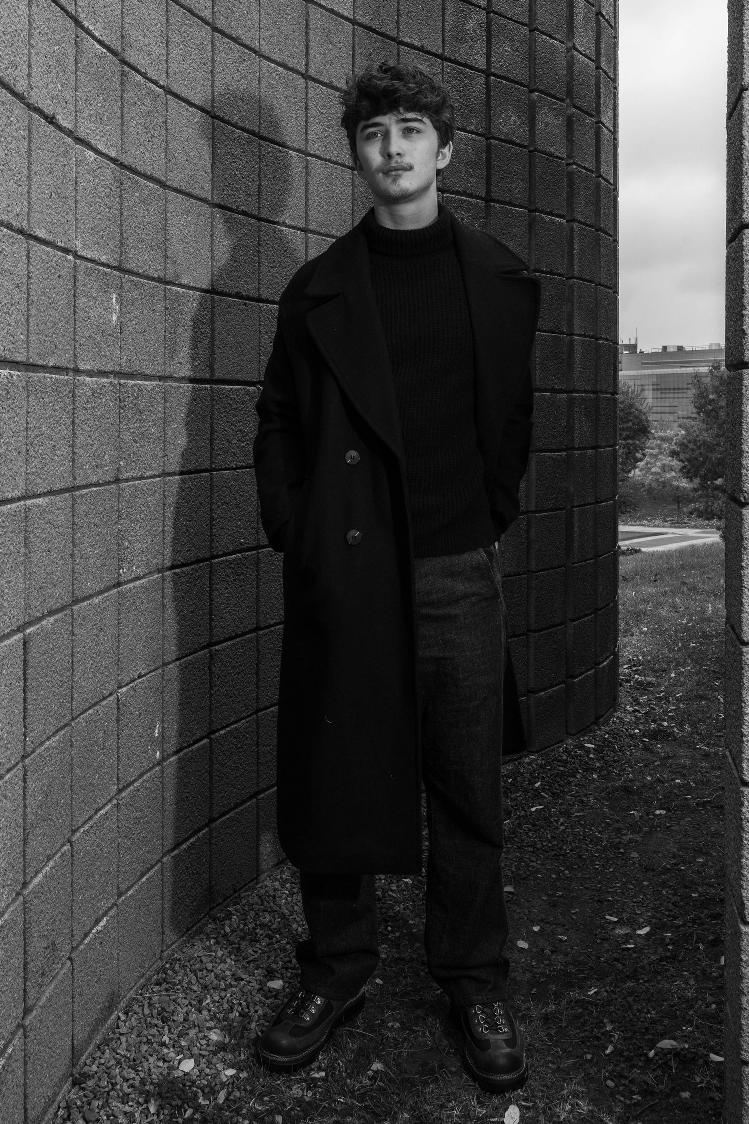Black and white portrait of a young man in a long dark coat and boots standing beside a curved brick wall casting a shadow.