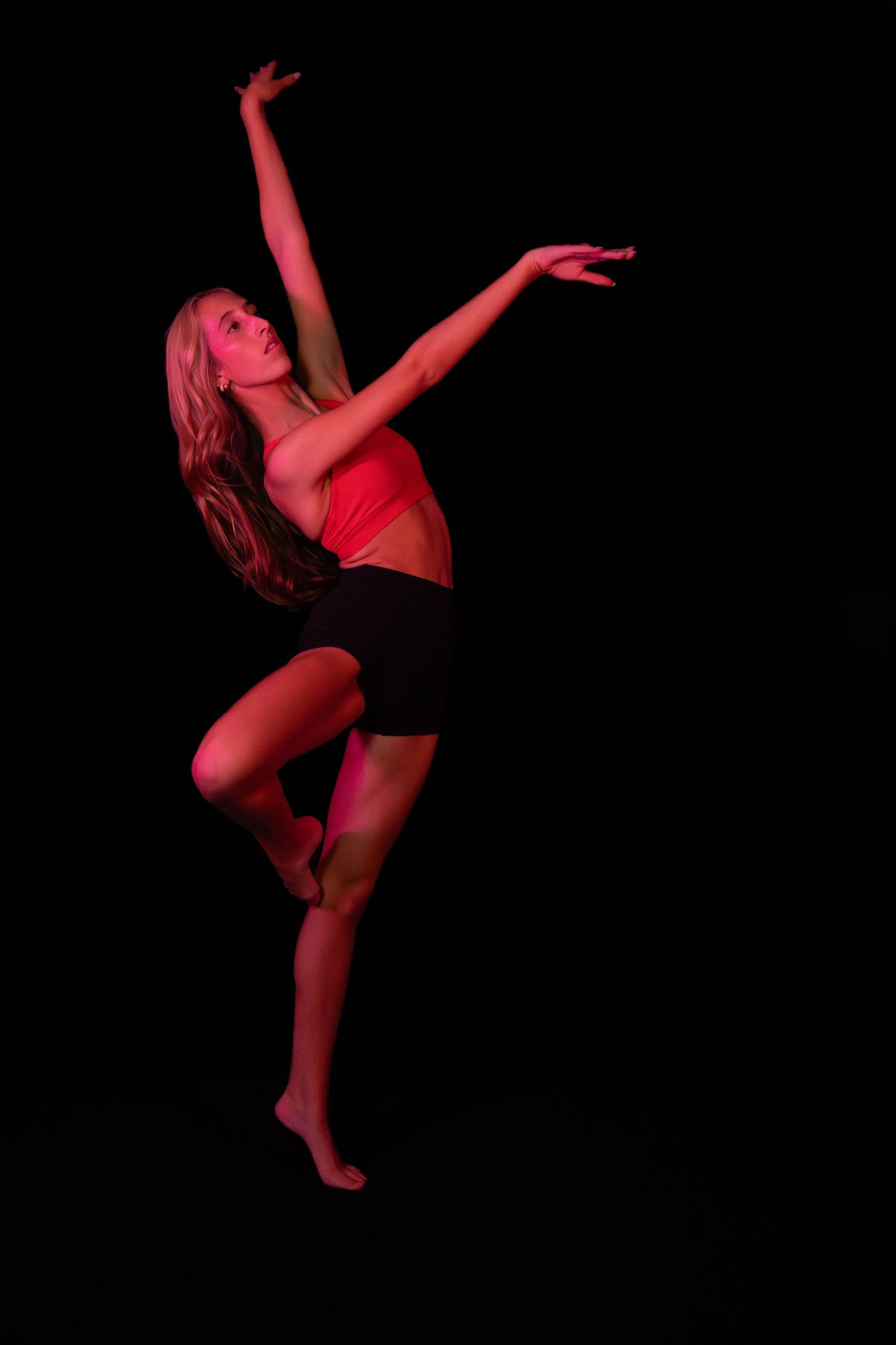 Dancer in a red top and black shorts balances on one leg with arms extended, lit by dramatic red light against a dark studio background.