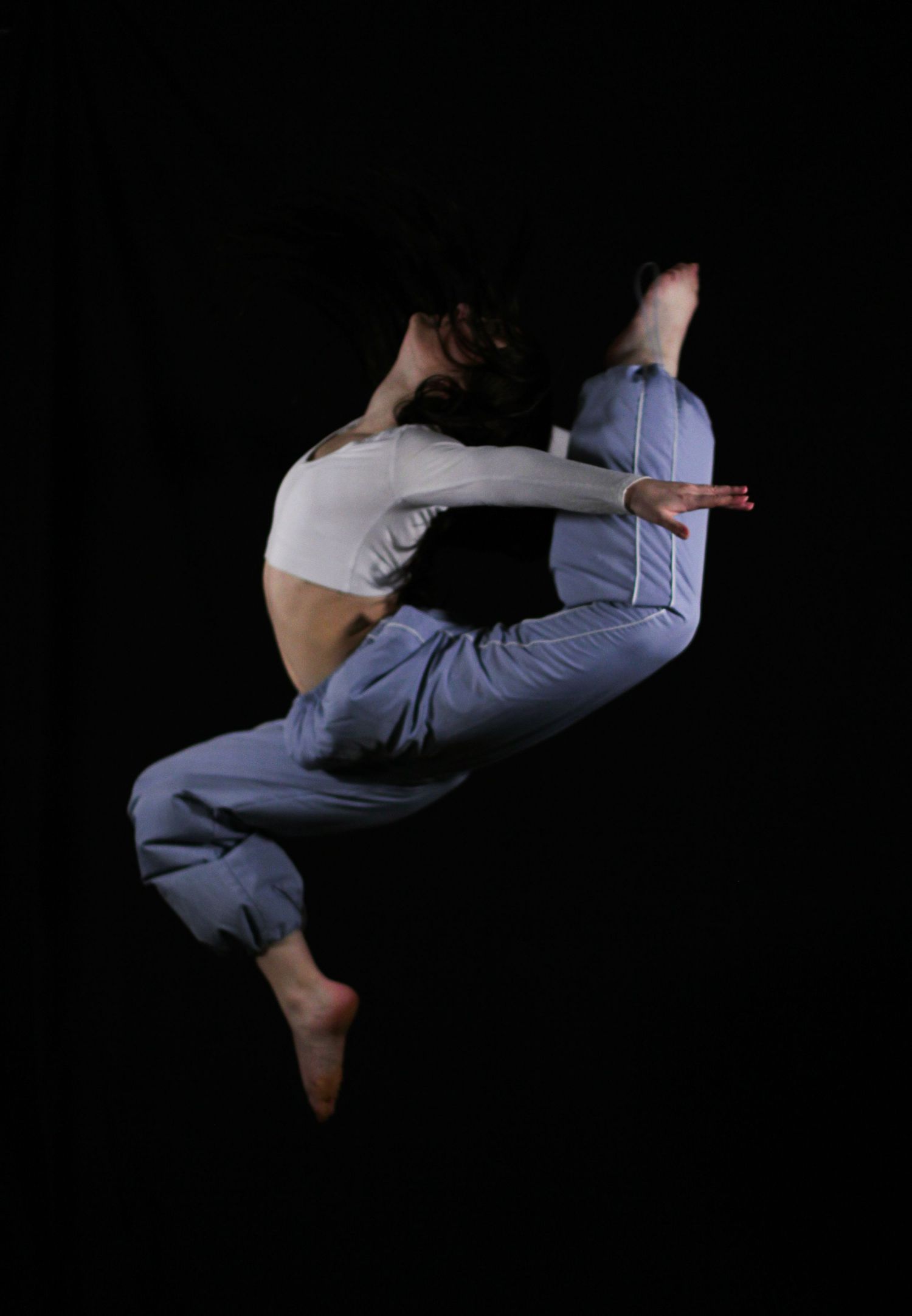 Dancer in a white crop top and loose blue pants leaps mid-air with arched back and bent legs against a dark black background.