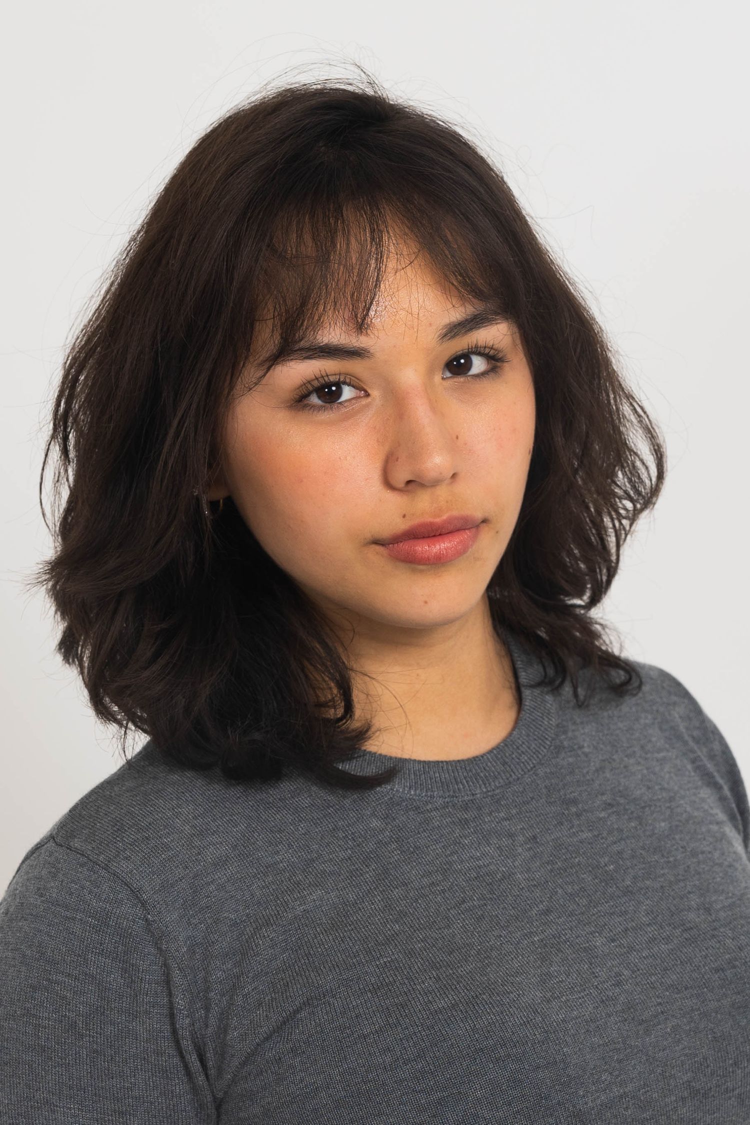 Portrait of a young woman with wavy dark shoulder-length hair and bangs, wearing a gray crewneck sweater against a plain light background.