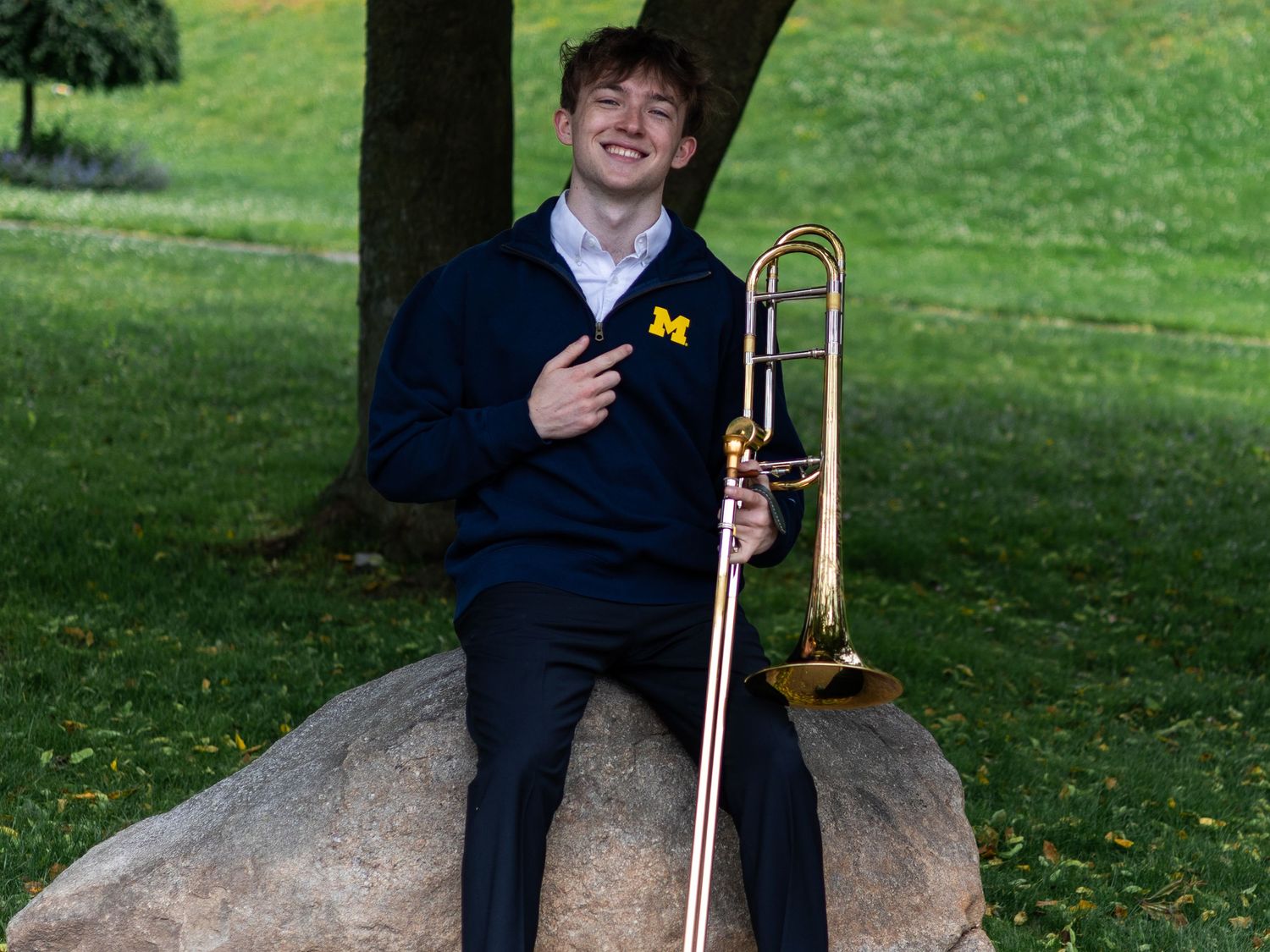 Smiling young man in University of Michigan hoodie holding a trombone outdoors on a rock.