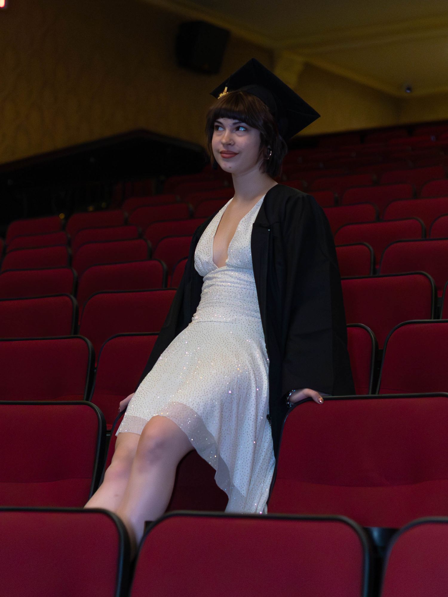 Young woman in white dress and graduation cap and gown posing in red theater seats.