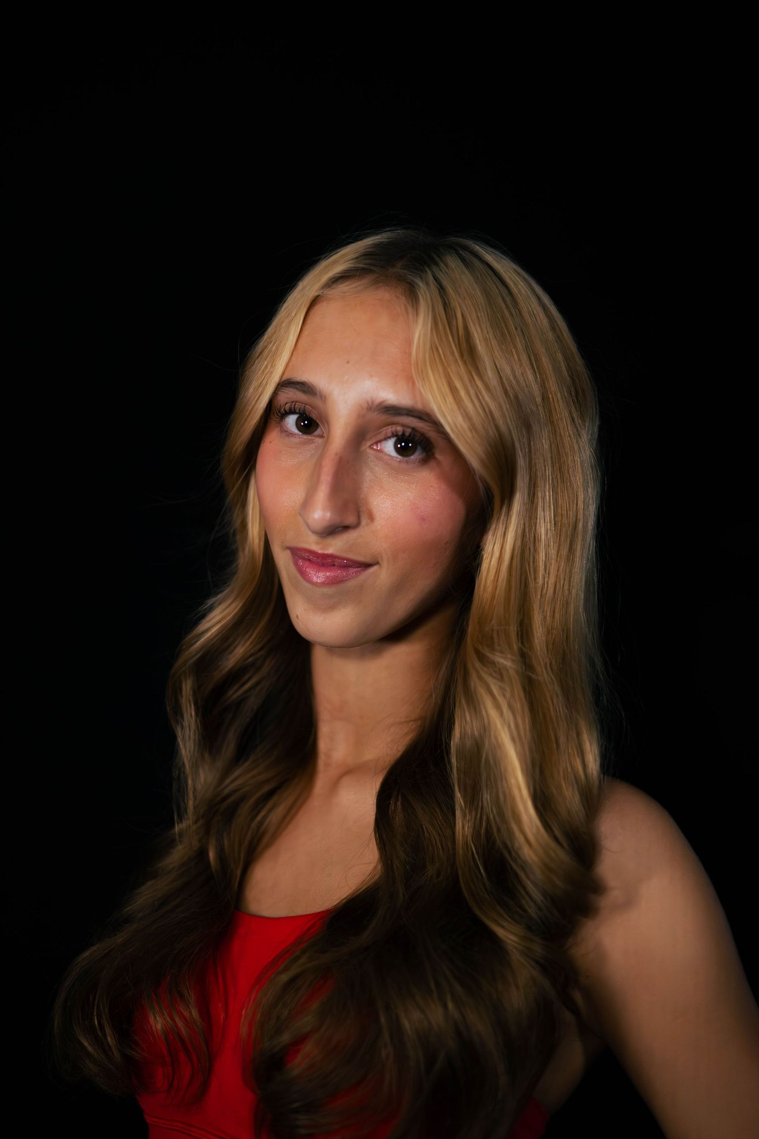 Portrait of a young woman with long wavy blonde and brown hair, wearing a red top and smiling softly against a black background.