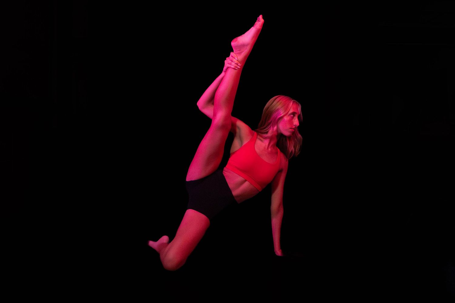 Dancer in red lighting performing a high vertical leg extension against a dark background, showcasing flexibility and athletic grace.