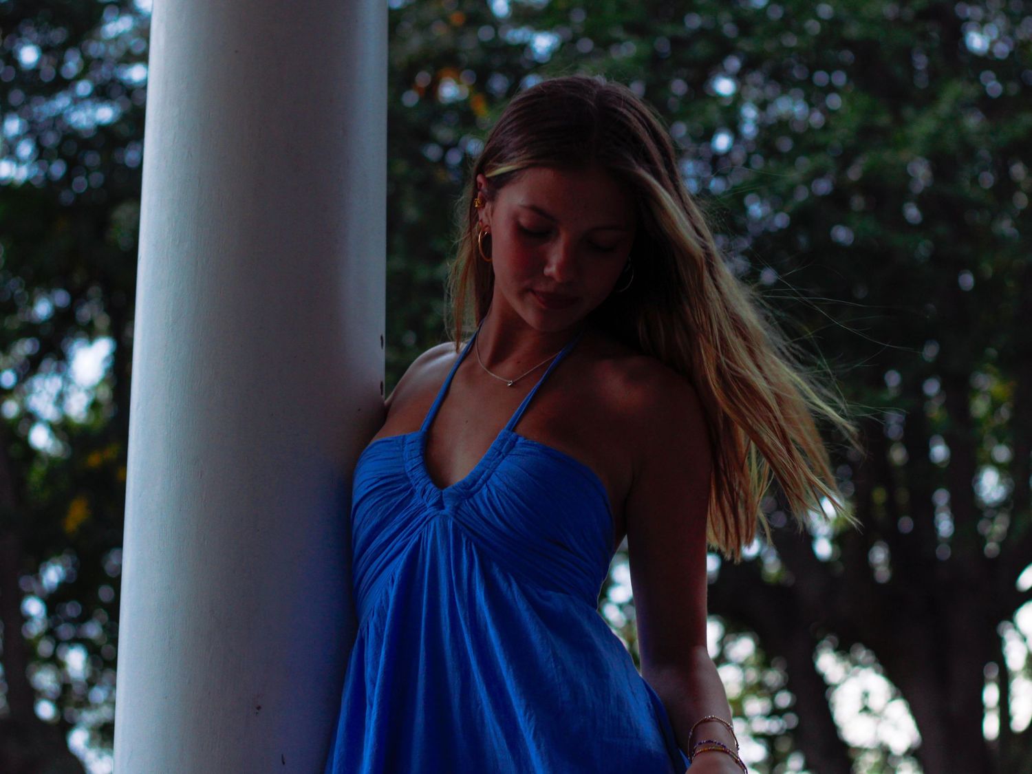 Young woman in a blue halter dress leans against a white column surrounded by lush trees.