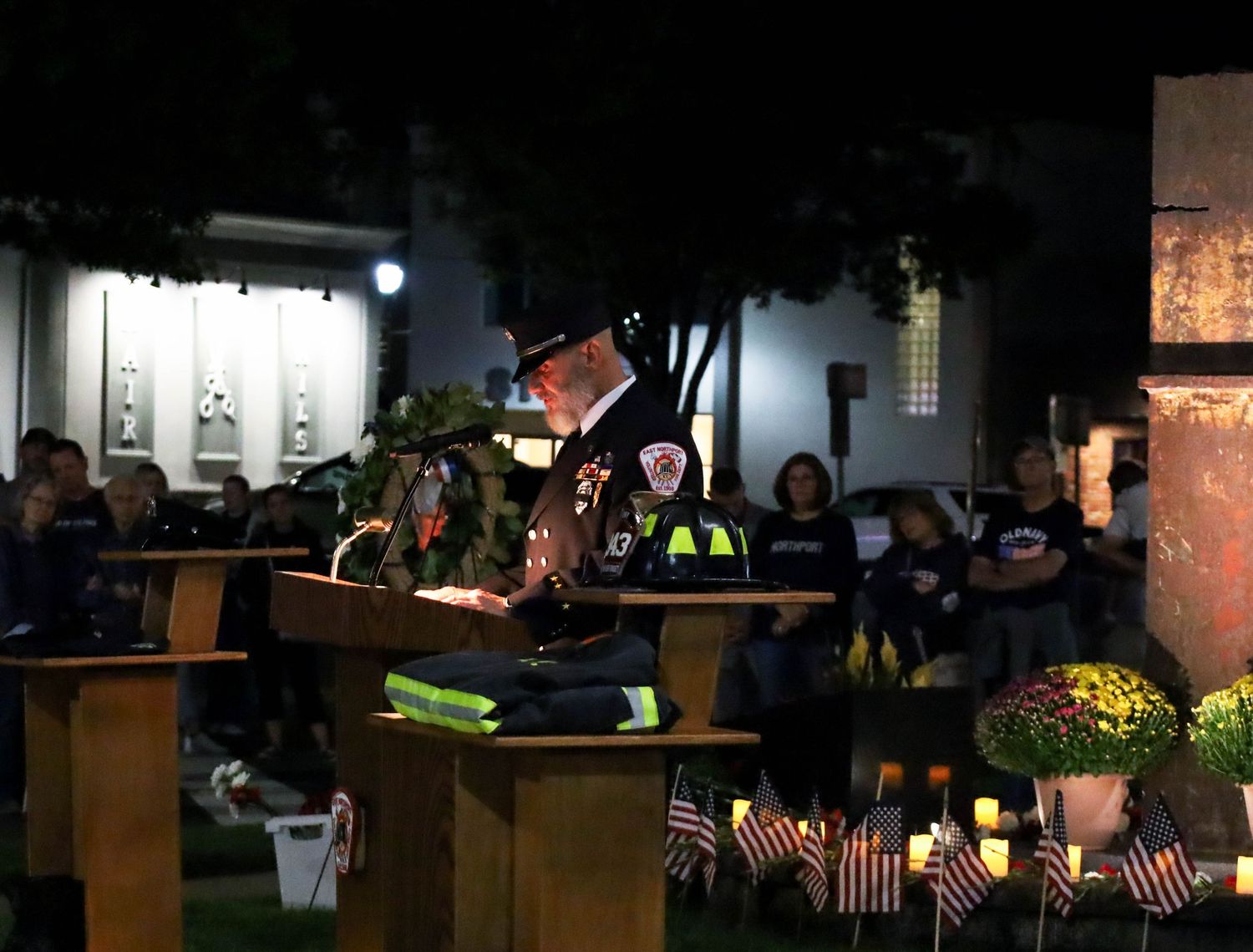 Firefighter speaks at a candlelit 9/11 memorial ceremony at night with American flags and attendees in background.