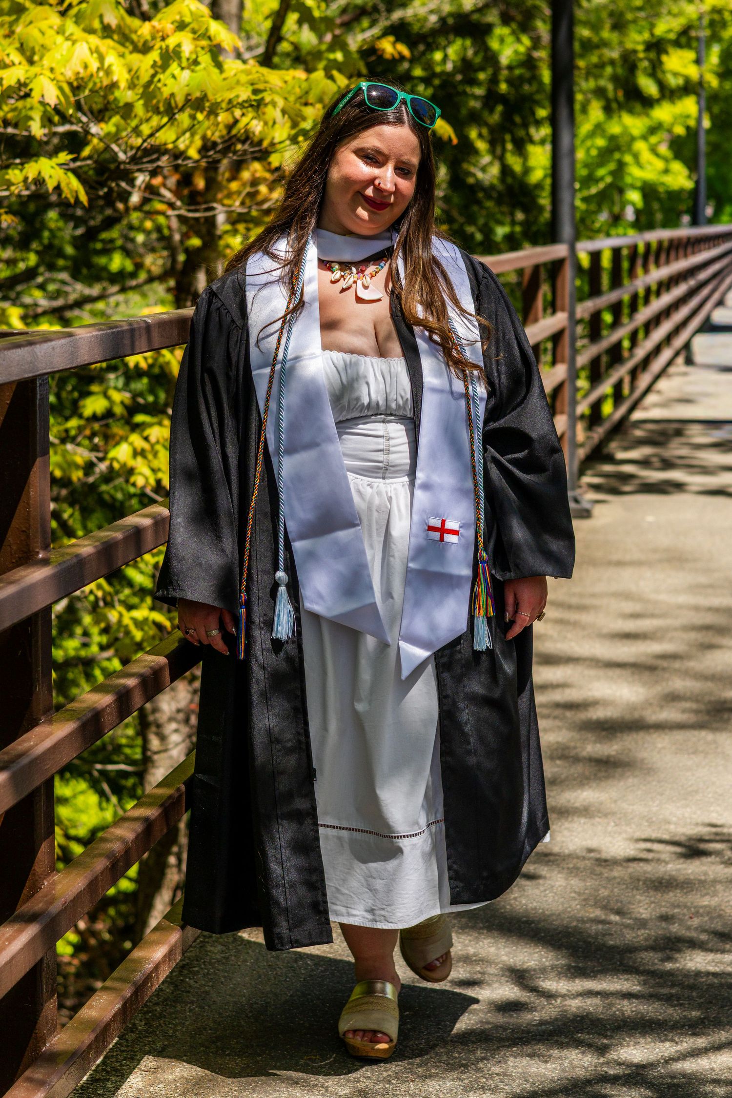 Young woman in black graduation gown and white stole walks on a wooden bridge surrounded by green trees.
