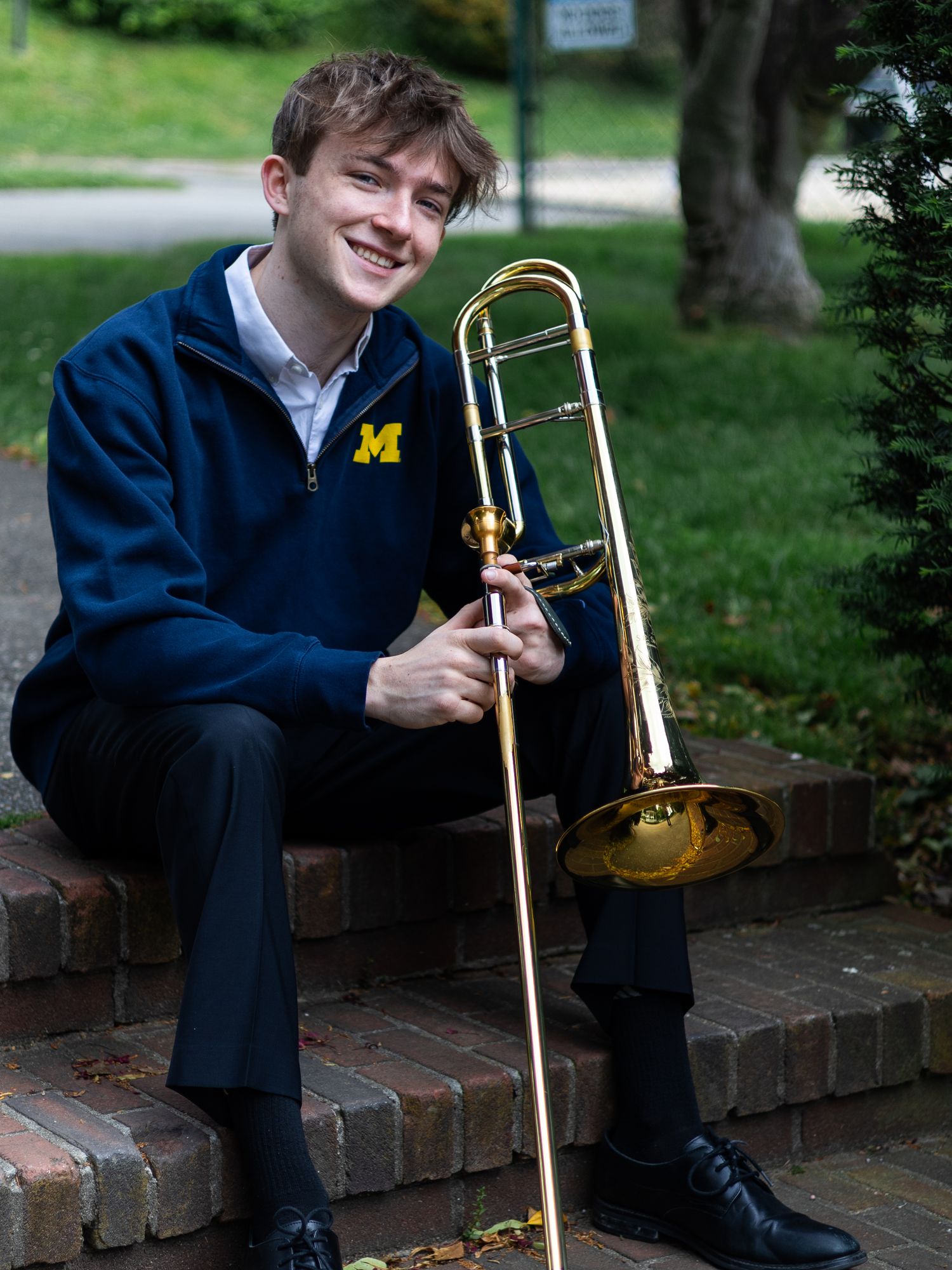 Young musician in University of Michigan jacket smiling while holding a trombone, seated on brick steps outdoors.