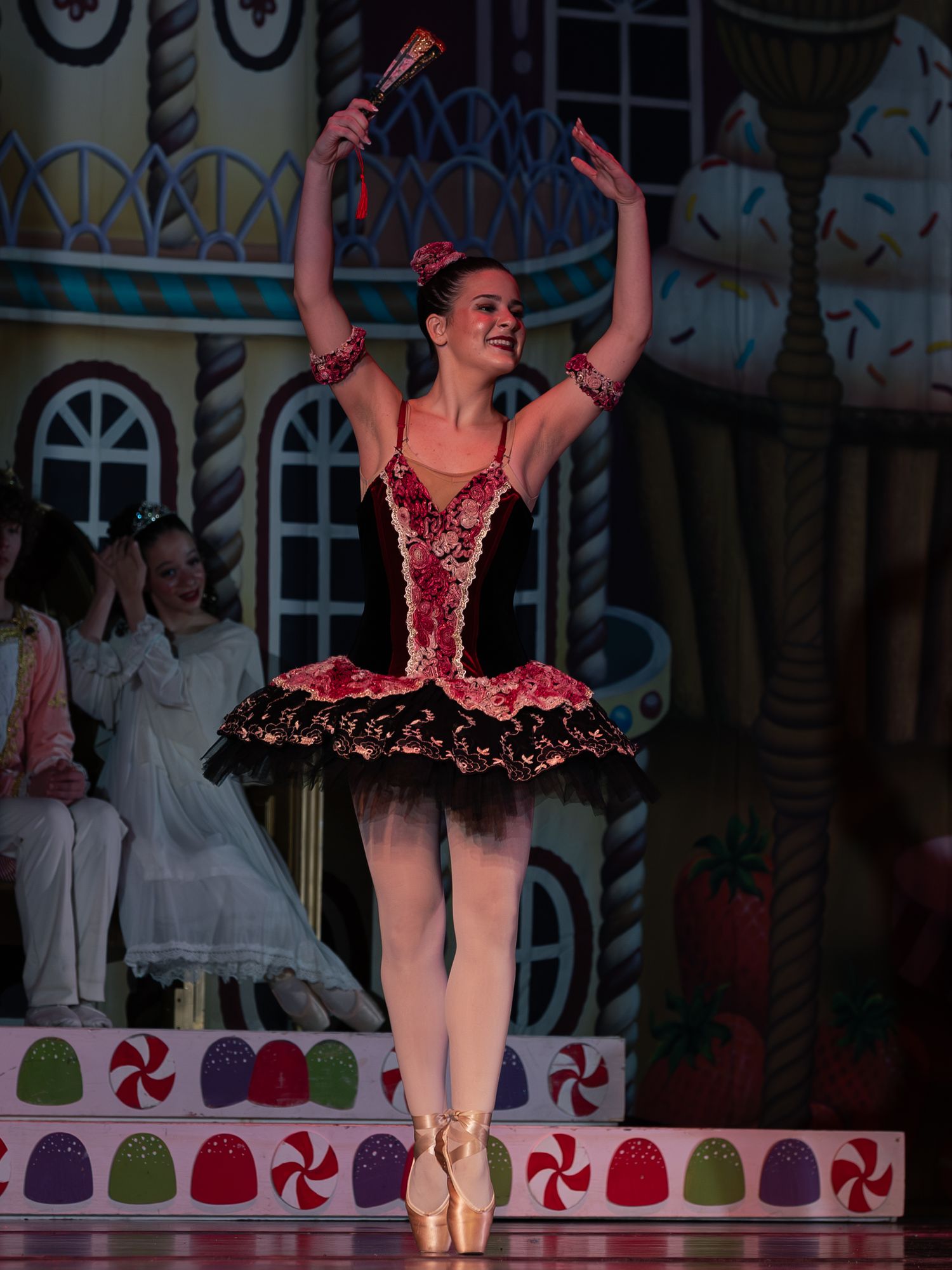 Ballerina in red and black tutu performs on pointe on a candy-themed Nutcracker stage set.