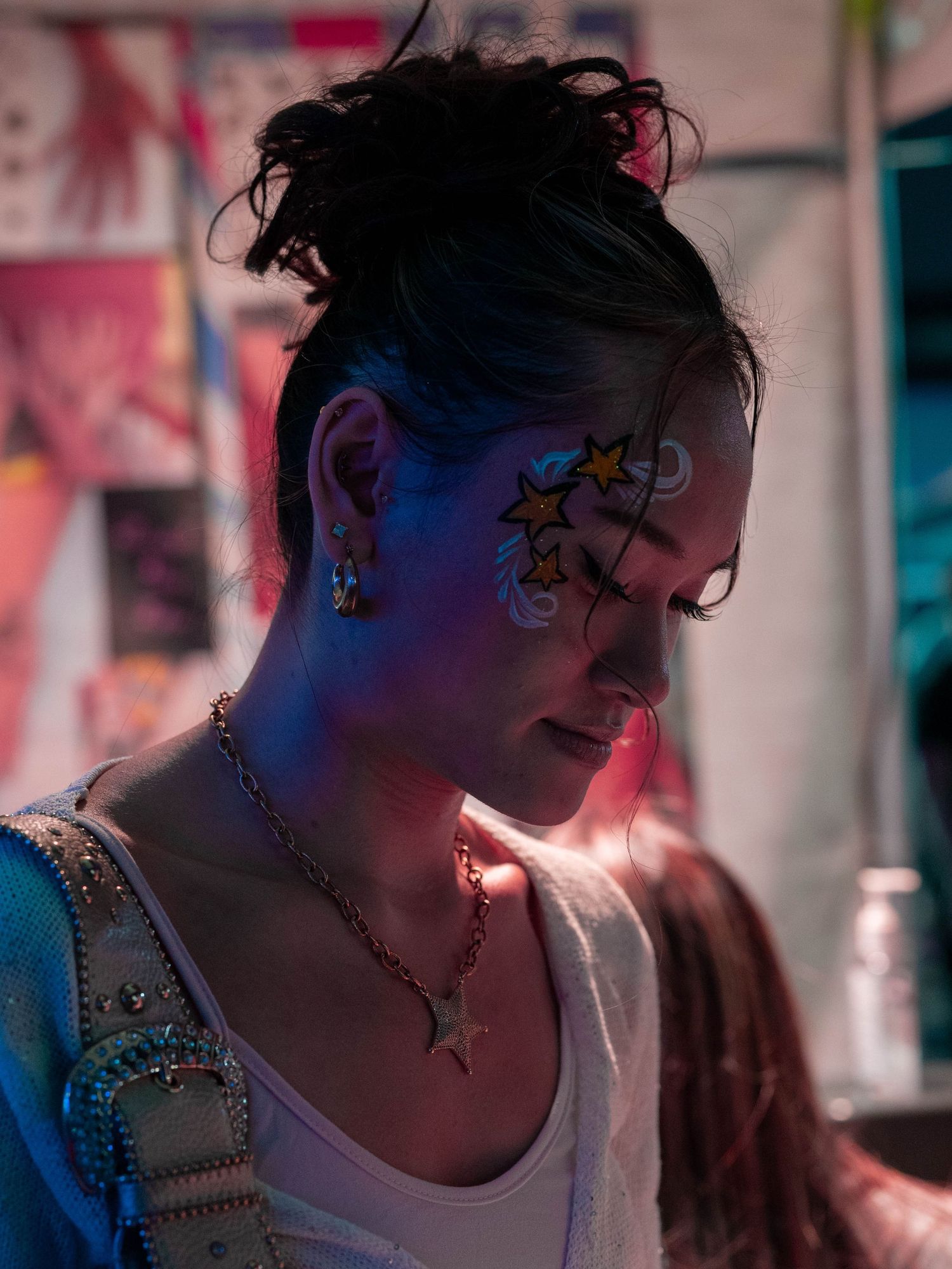 Young woman with star face paint and star necklace under colorful festival lighting.