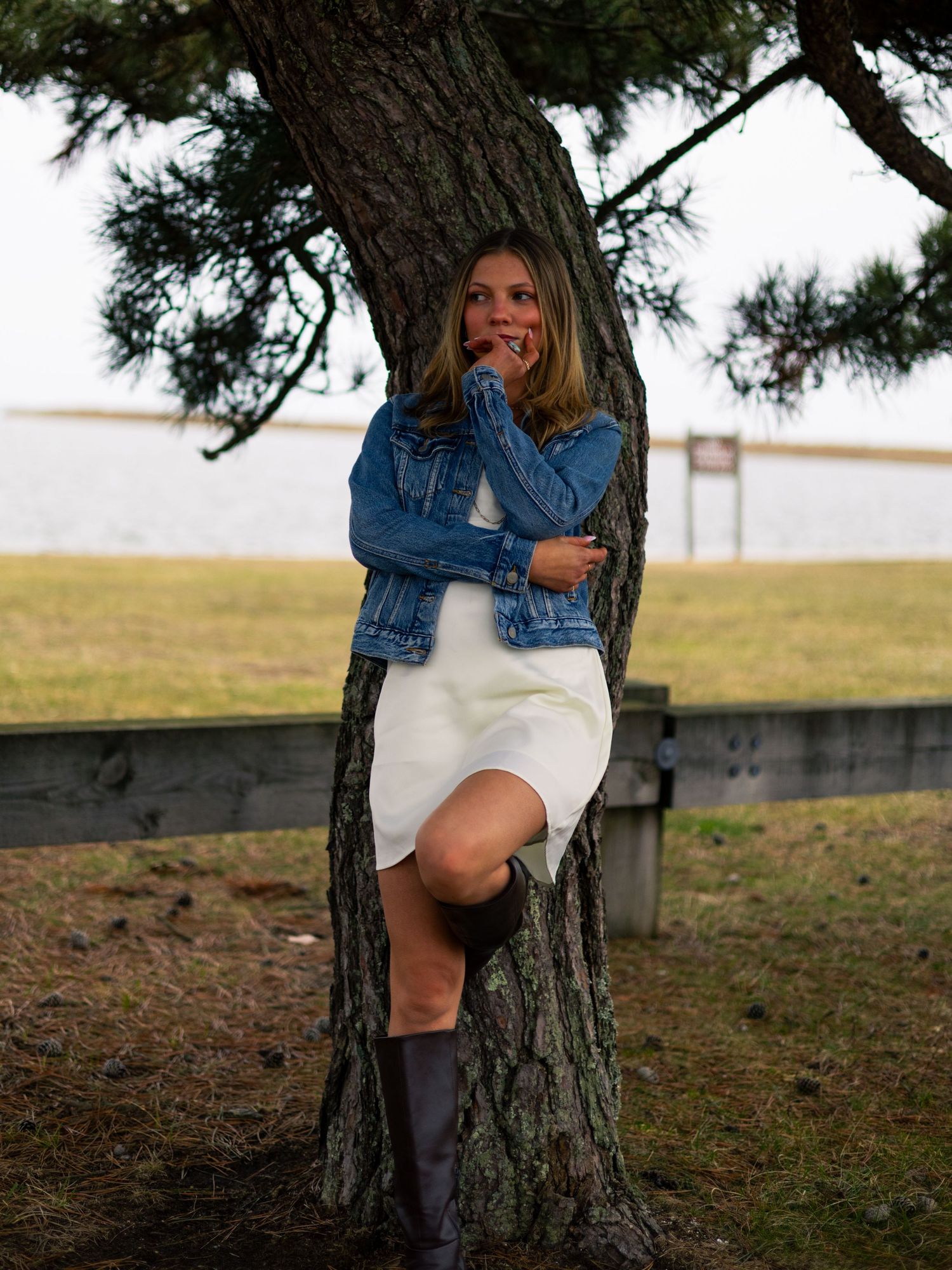 Woman in a denim jacket, white dress and tall brown boots leans against a pine tree with a grassy field and water behind her.