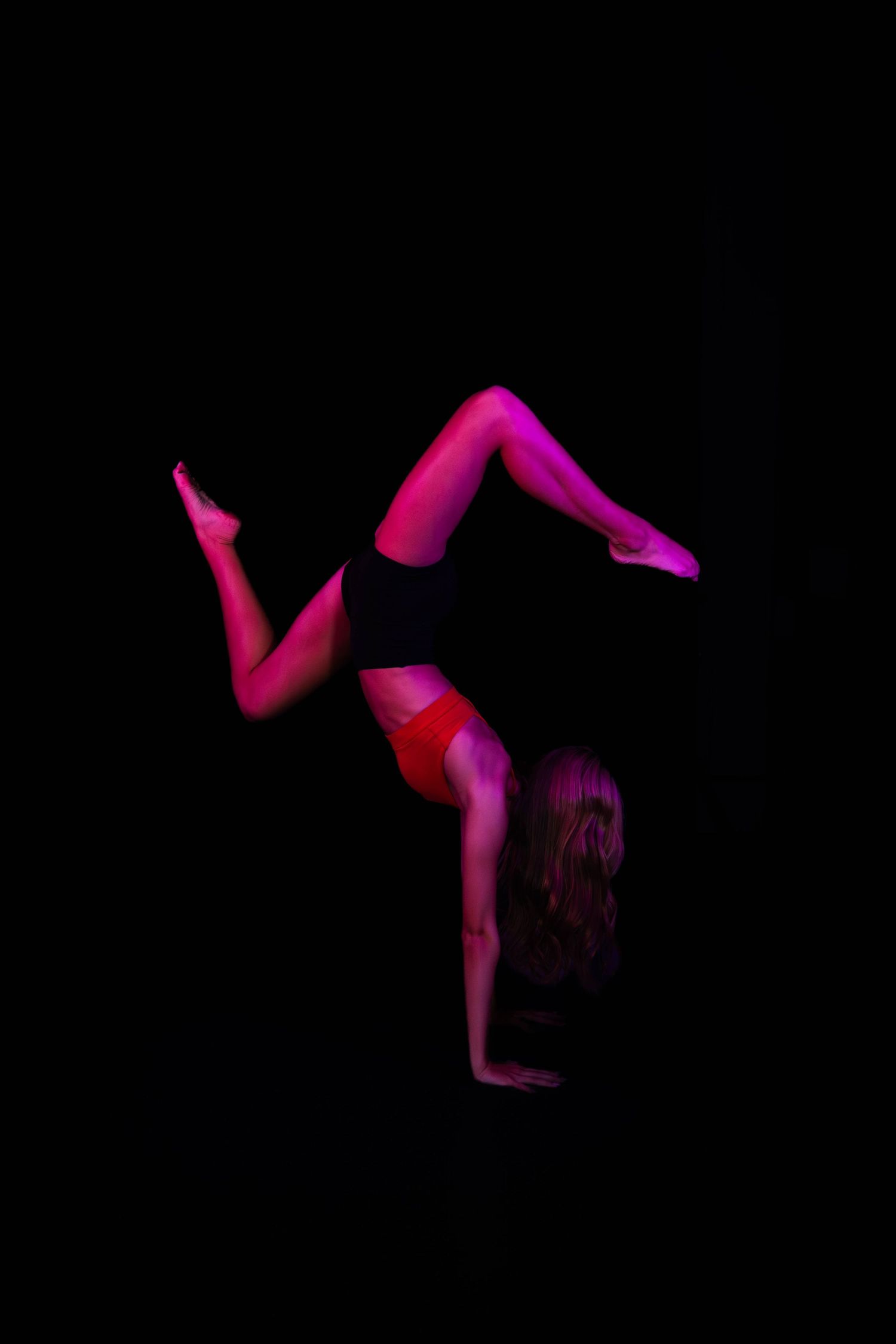 Dancer in red and black athletic wear performs a handstand with legs split, lit by pink stage lighting against a dark background.