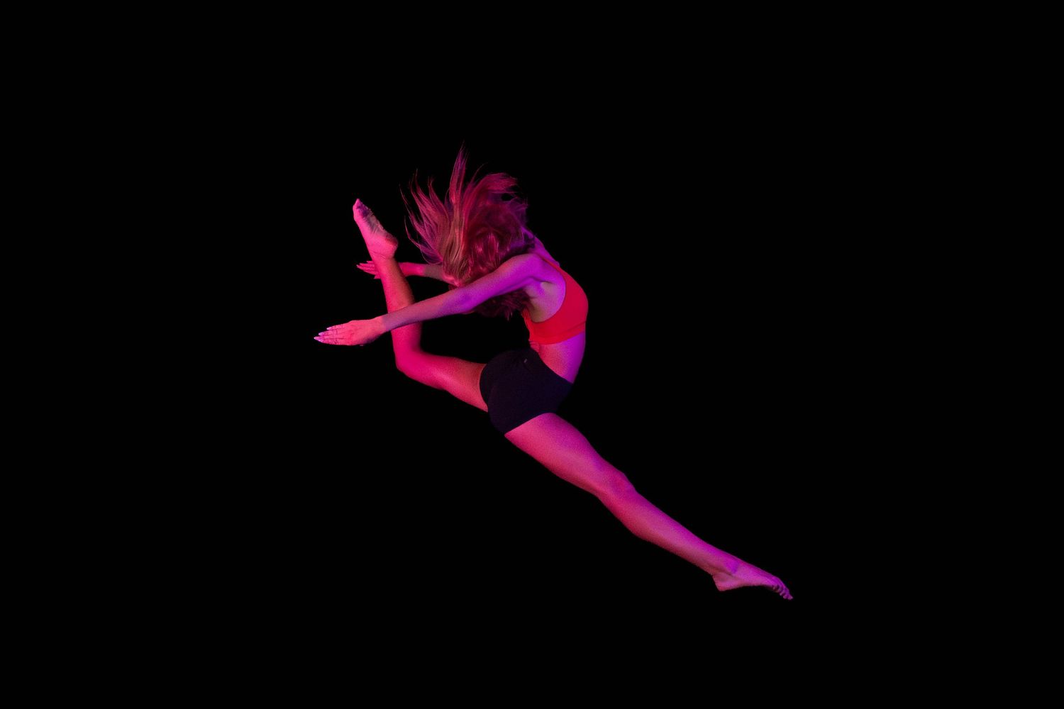 Dancer leaps mid-air in a split jump against a black background, bathed in vibrant pink and magenta stage lighting.