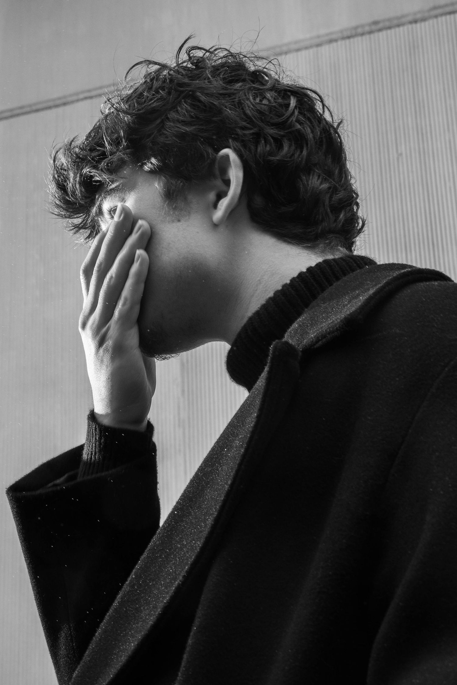 Black and white profile portrait of a curly-haired man in a dark coat and turtleneck covering his face with one hand.