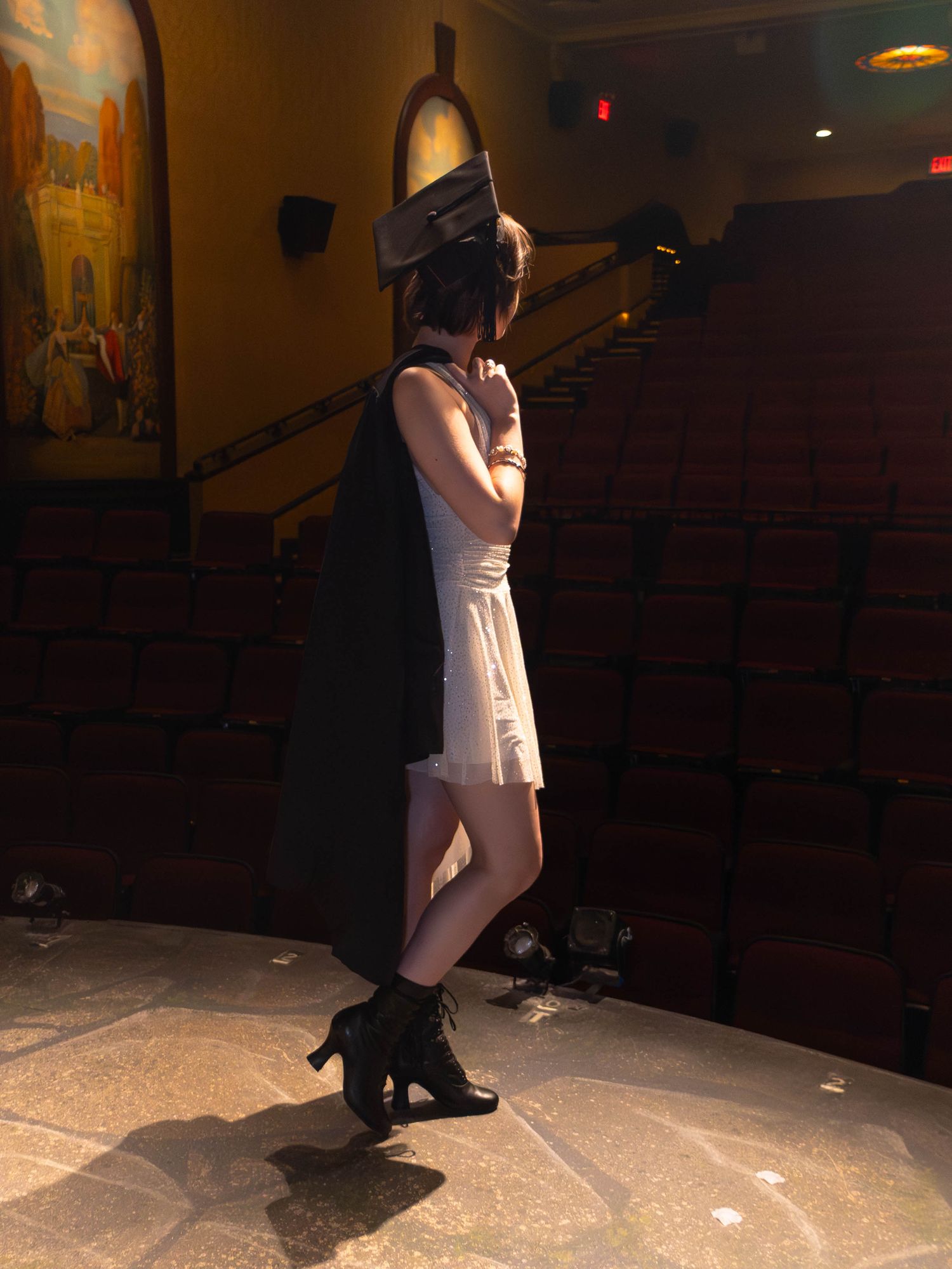 Woman in white dress, black cape, and heeled boots poses in an ornate theater with warm lighting and stadium seating.
