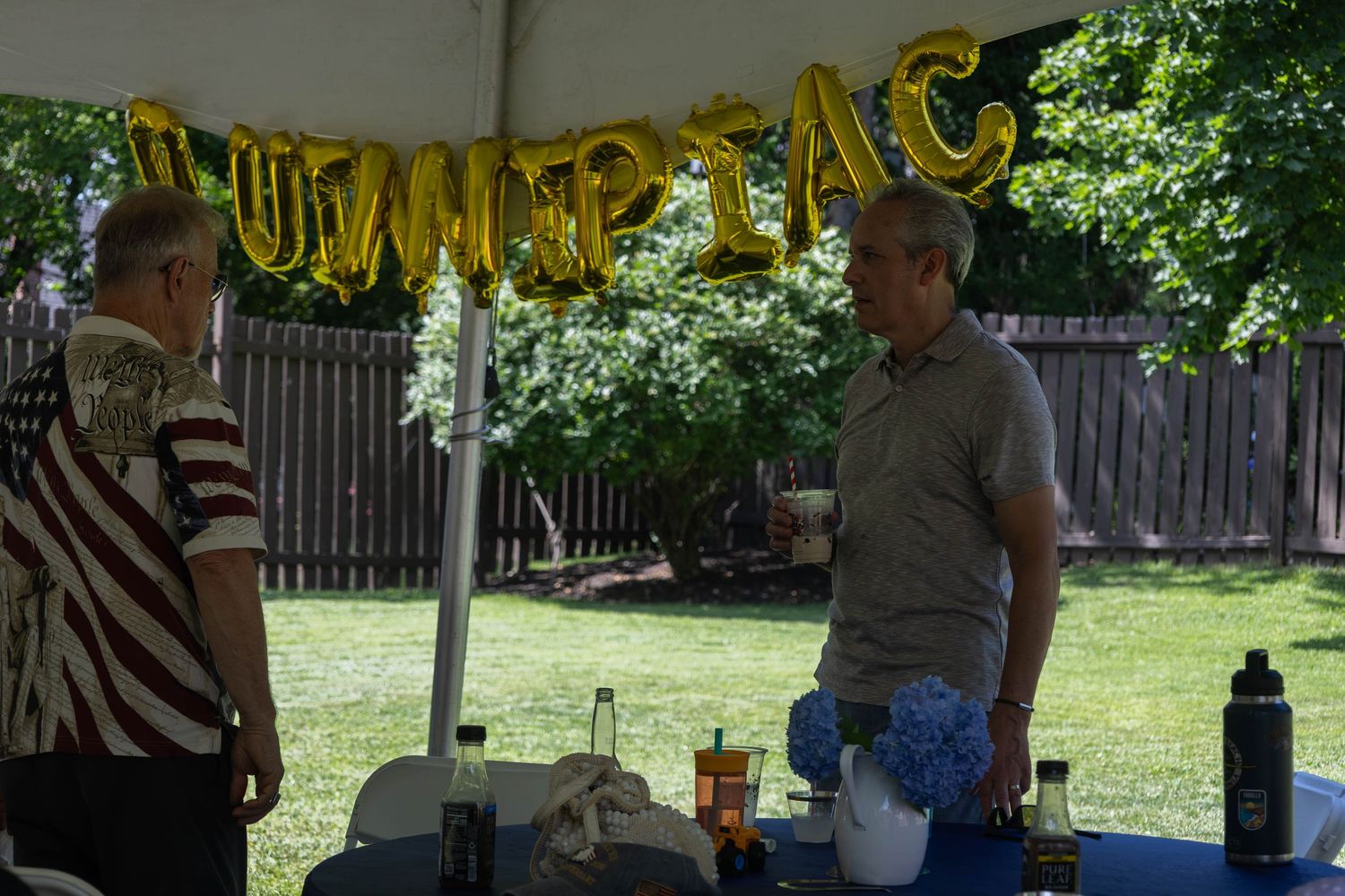 Two men talking under a tent decorated with gold QUINNIPIAC balloon letters at a graduation party.