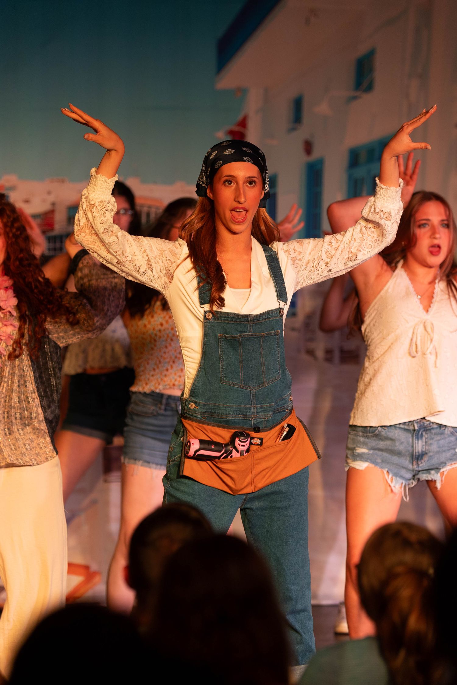 Young woman in denim overalls and headband performs energetically on stage in a Greek-themed musical production.