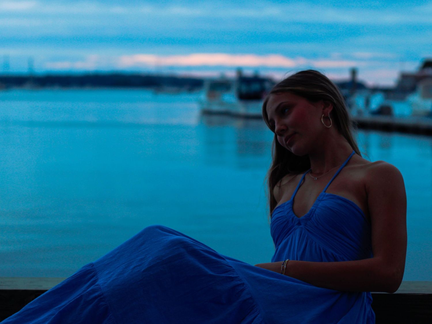 Woman in blue halter maxi dress sitting by waterfront at dusk with soft blue sky.