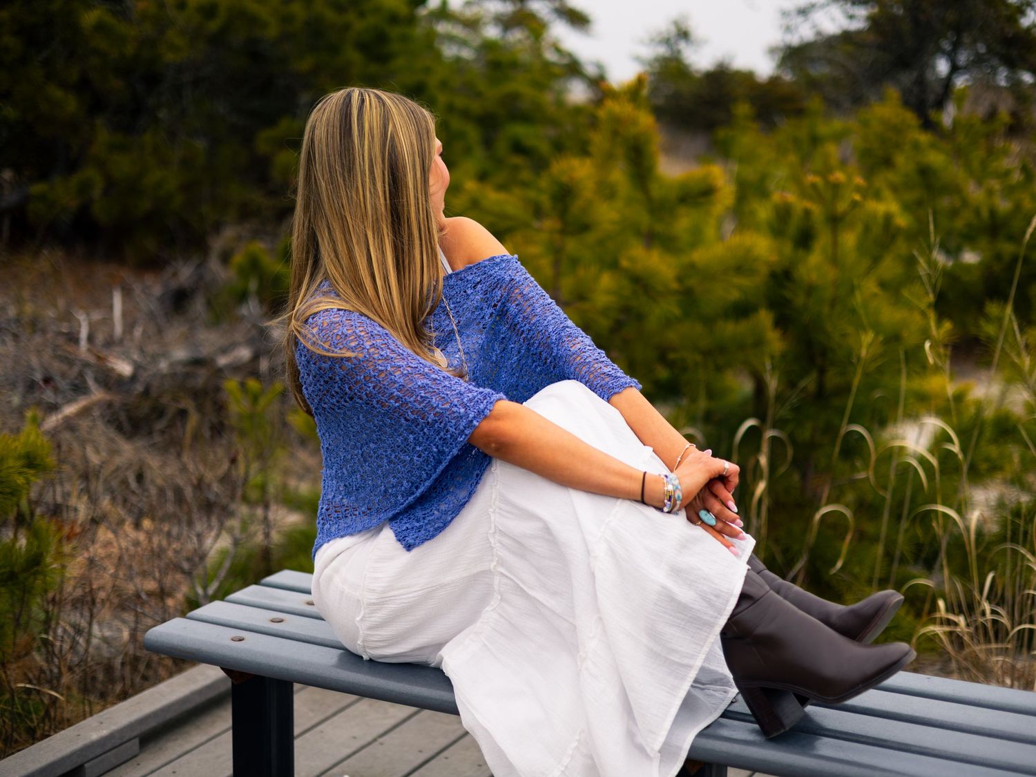 Woman in blue sweater and white skirt sitting on bench, gazing at lush green landscape outdoors.
