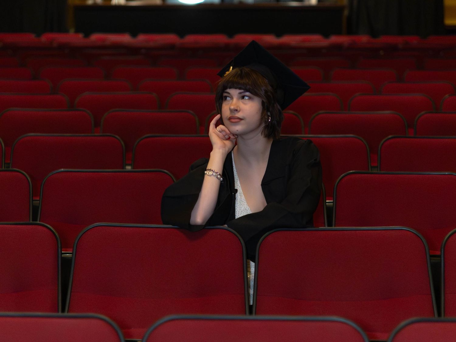 A graduate in cap and gown sits alone among red theater seats, gazing thoughtfully upward.