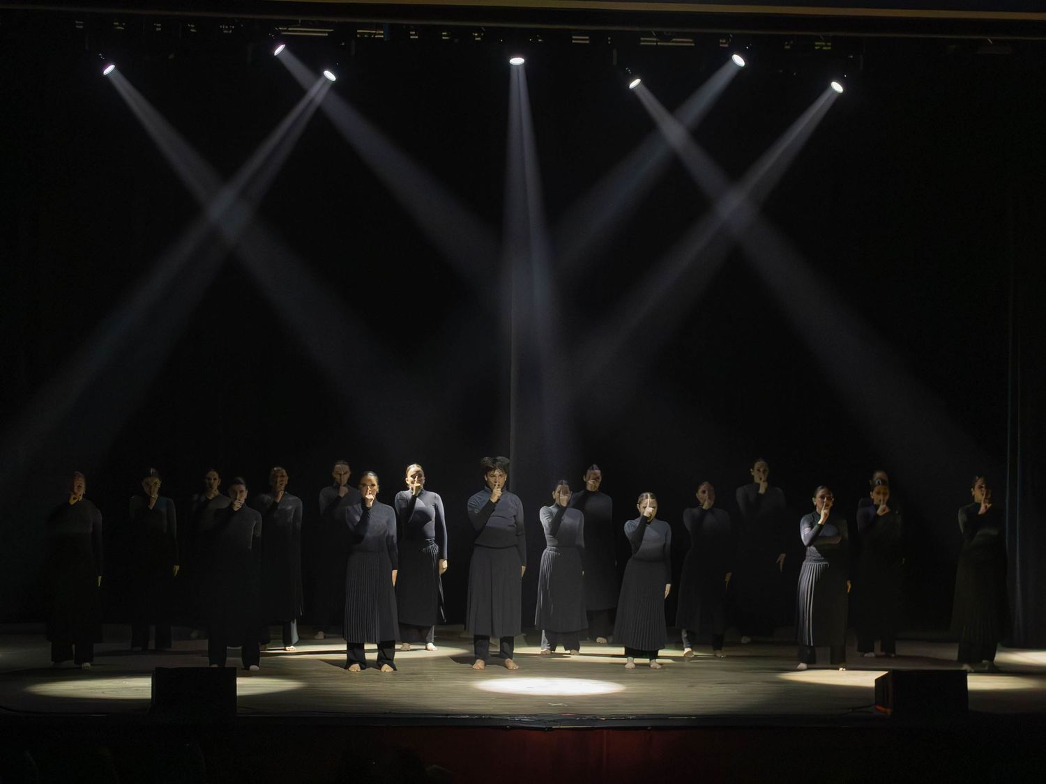 A choir in black robes performs on a dramatic stage under bright spotlights in a dark theater setting.
