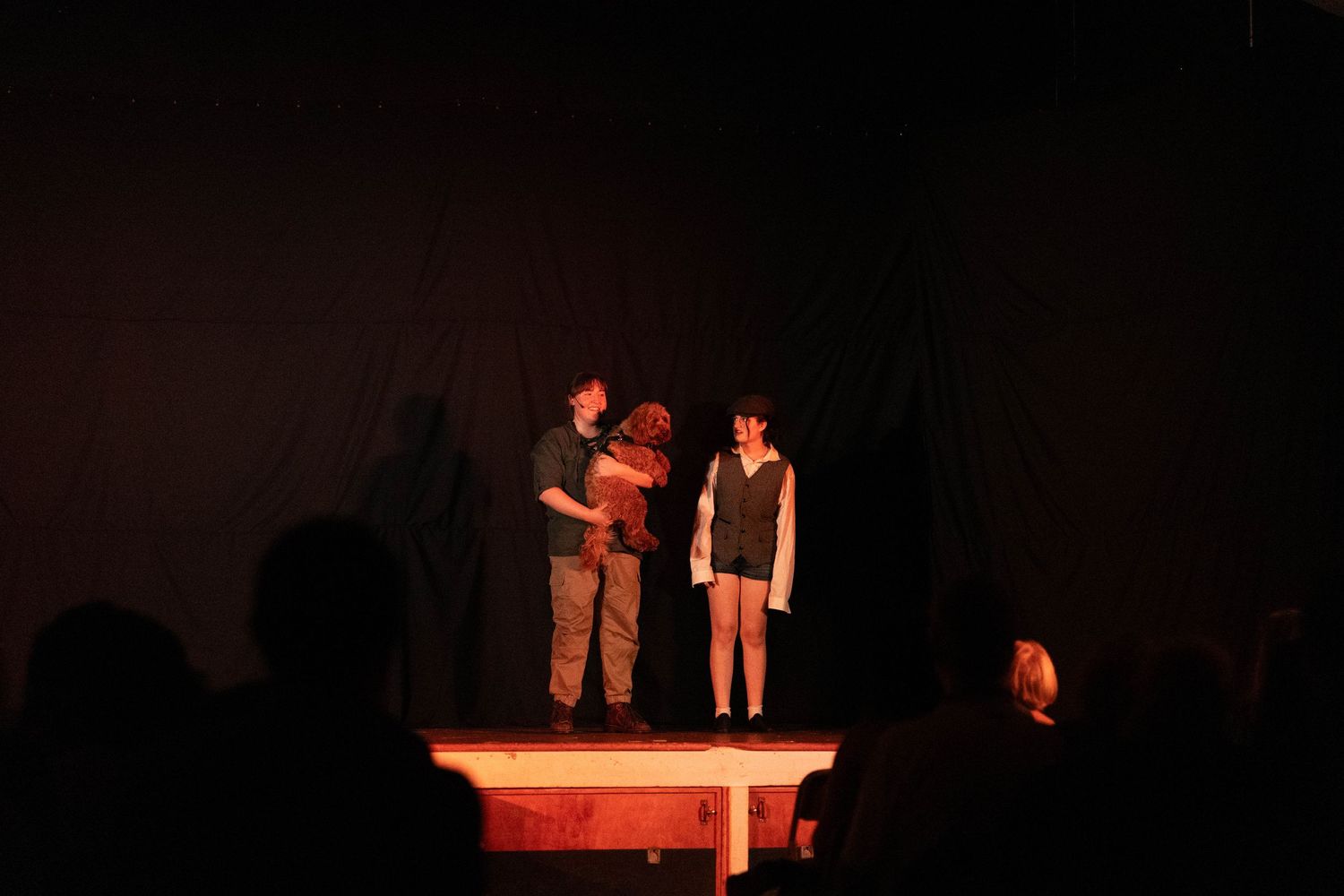 Three performers on a dimly lit stage during a theater production, with an audience visible in the foreground.