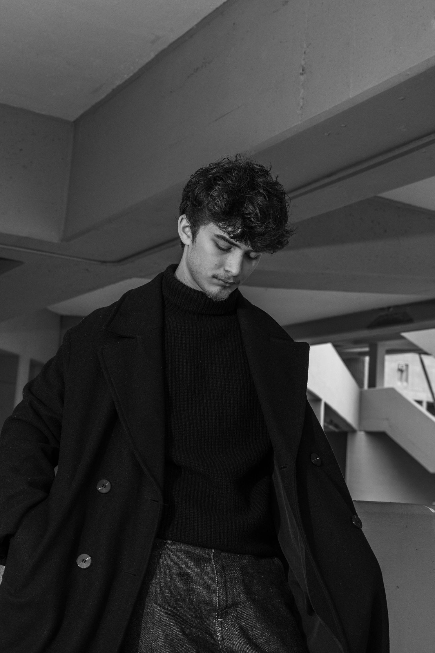 Black and white portrait of a young man with curly dark hair in a long wool coat and turtleneck, looking down inside a concrete building.