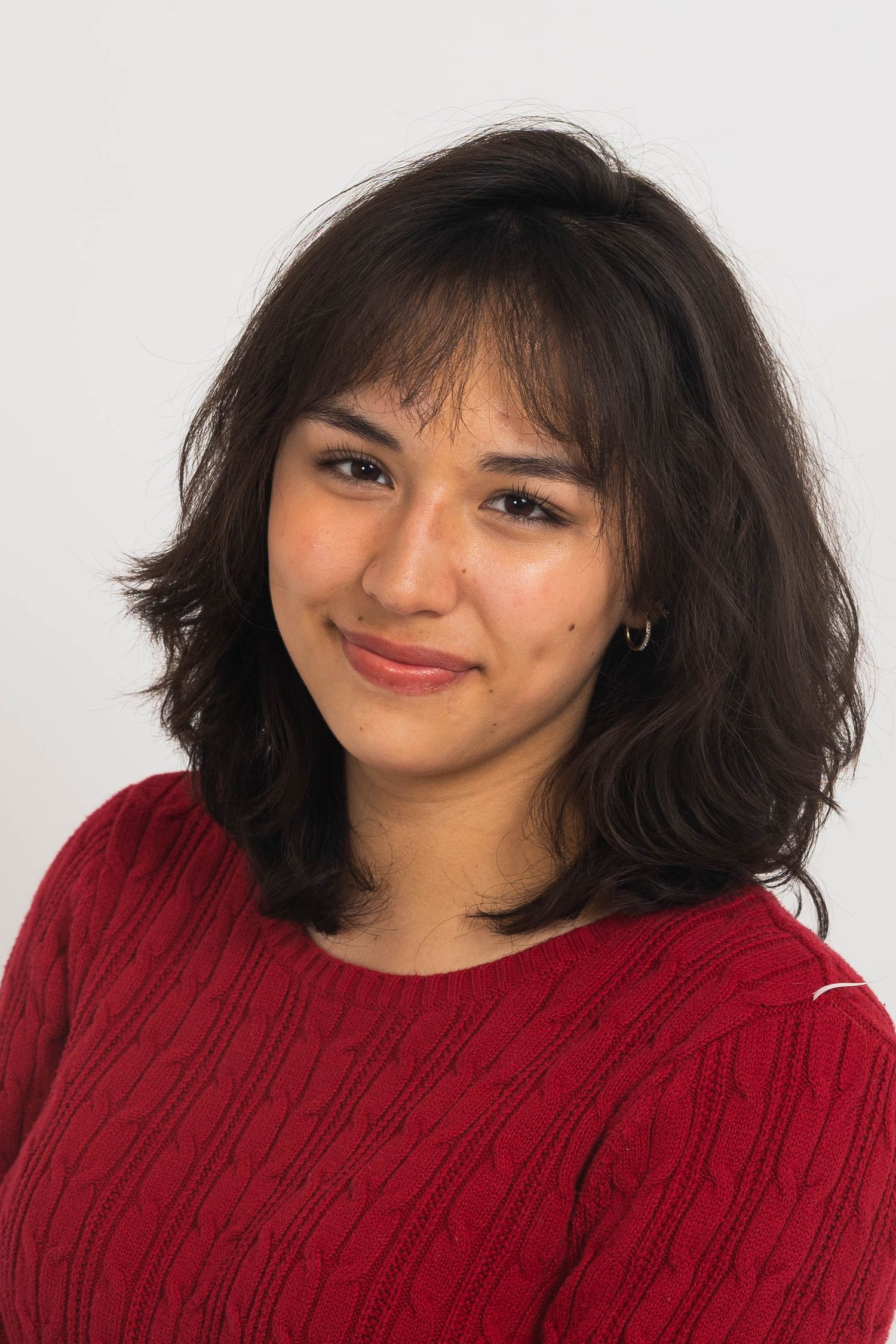 Smiling young woman with shoulder-length wavy dark hair and bangs wearing a red cable-knit sweater and small gold hoop earring.
