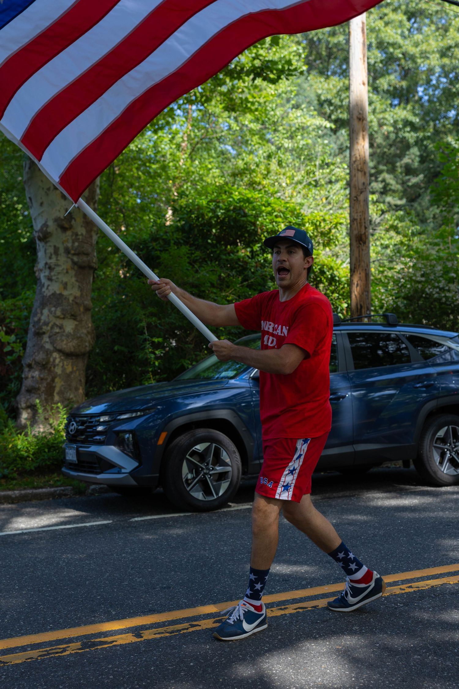 Man in red outfit energetically carries large American flag along tree-lined road during patriotic parade.