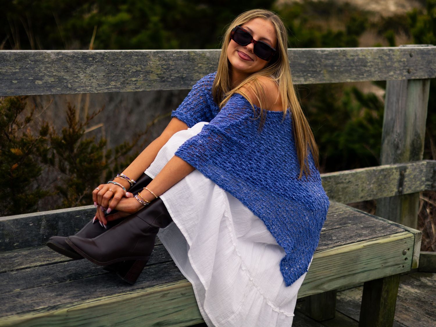 Woman in sunglasses, a blue knit top, white skirt and gray suede boots sits smiling on a weathered wooden bench near coastal dunes.