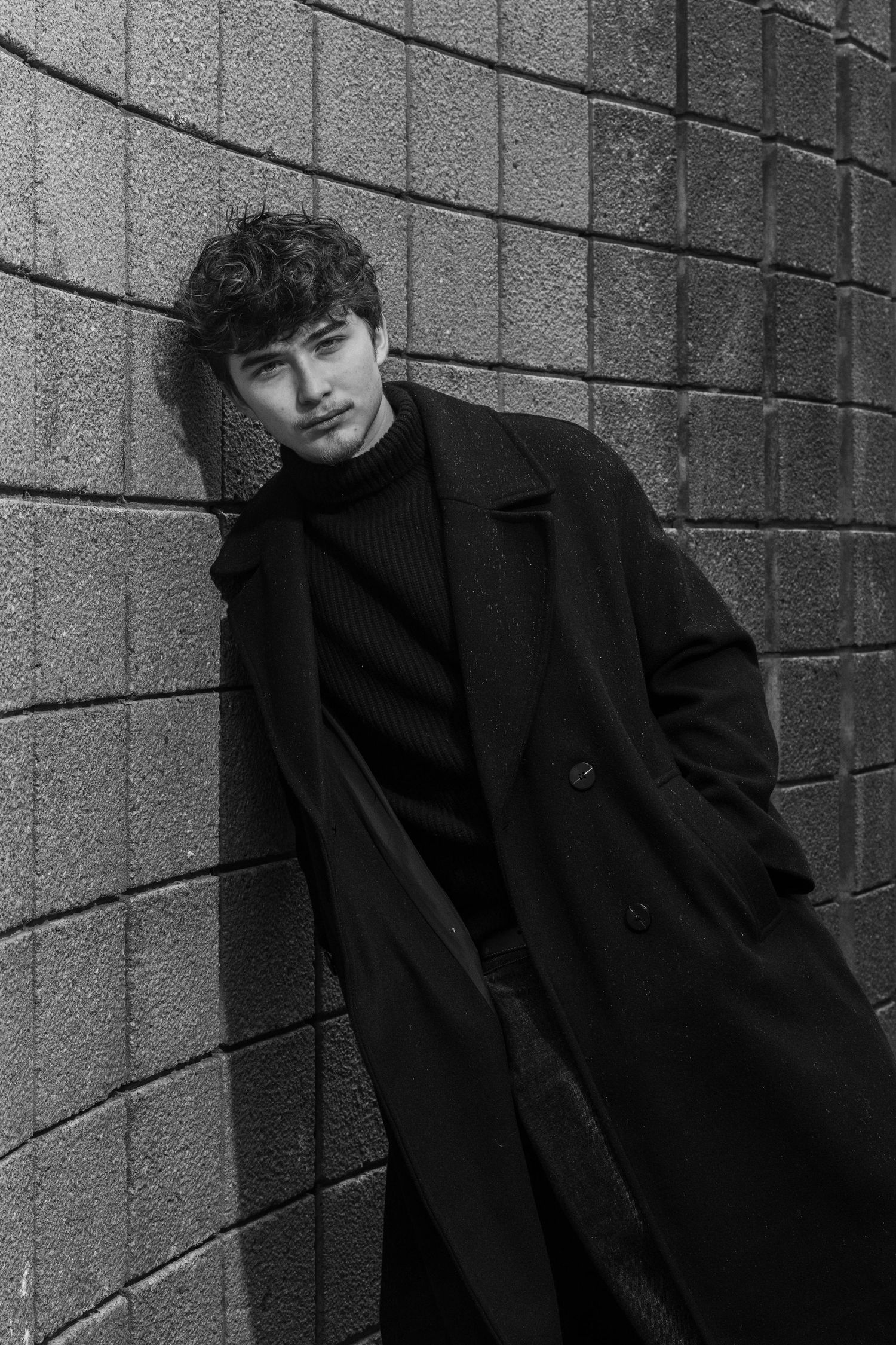 Black and white portrait of a young man with curly hair leaning against a curved brick wall in a long dark overcoat and turtleneck.