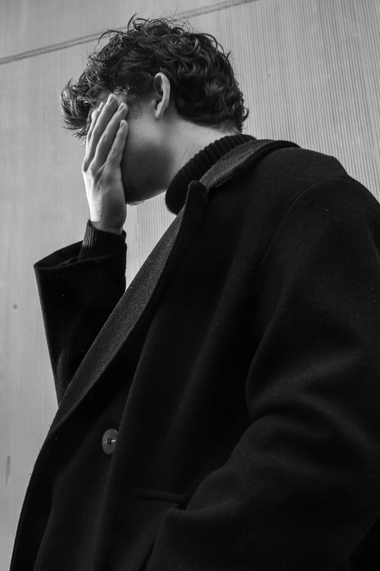 Black and white photo of a curly-haired man in a dark wool coat and turtleneck covering his face with one hand against a paneled wall.