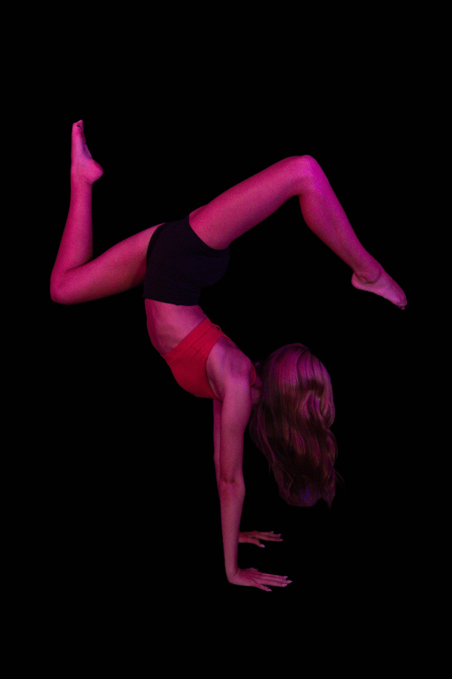 Gymnast in a handstand with legs split in mid-air, lit by dramatic pink and magenta stage light against a black background.