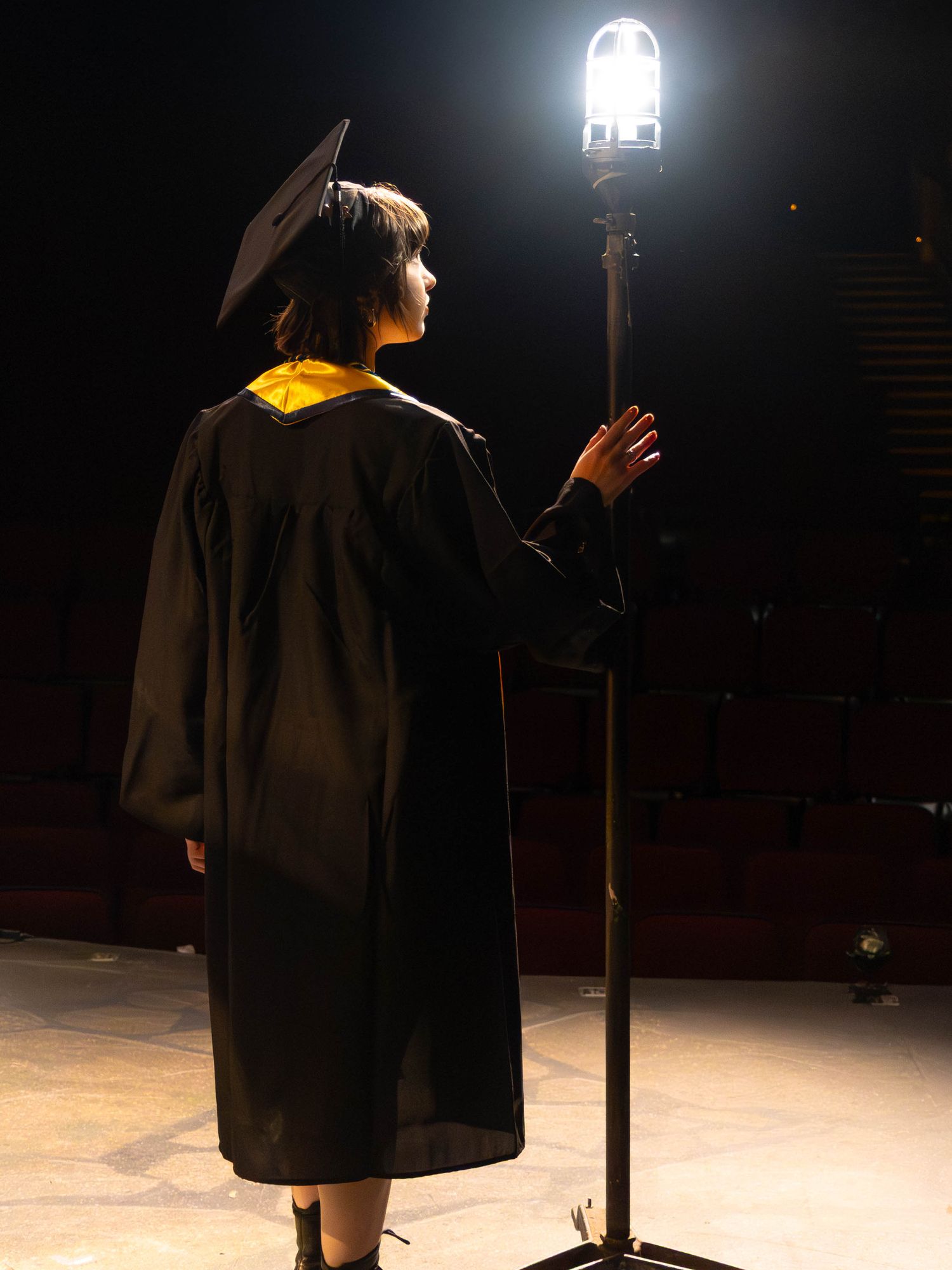 Graduate in black cap and gown stands at illuminated microphone stand on dark stage during commencement ceremony.