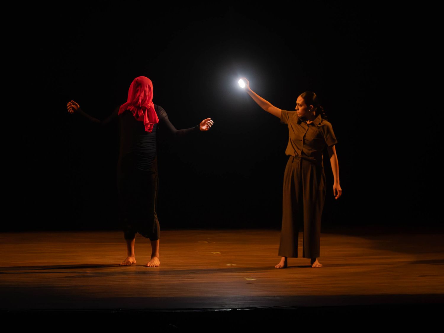 Two performers on a dark stage, one with red hair and one reaching toward a bright light in a dramatic dance performance.