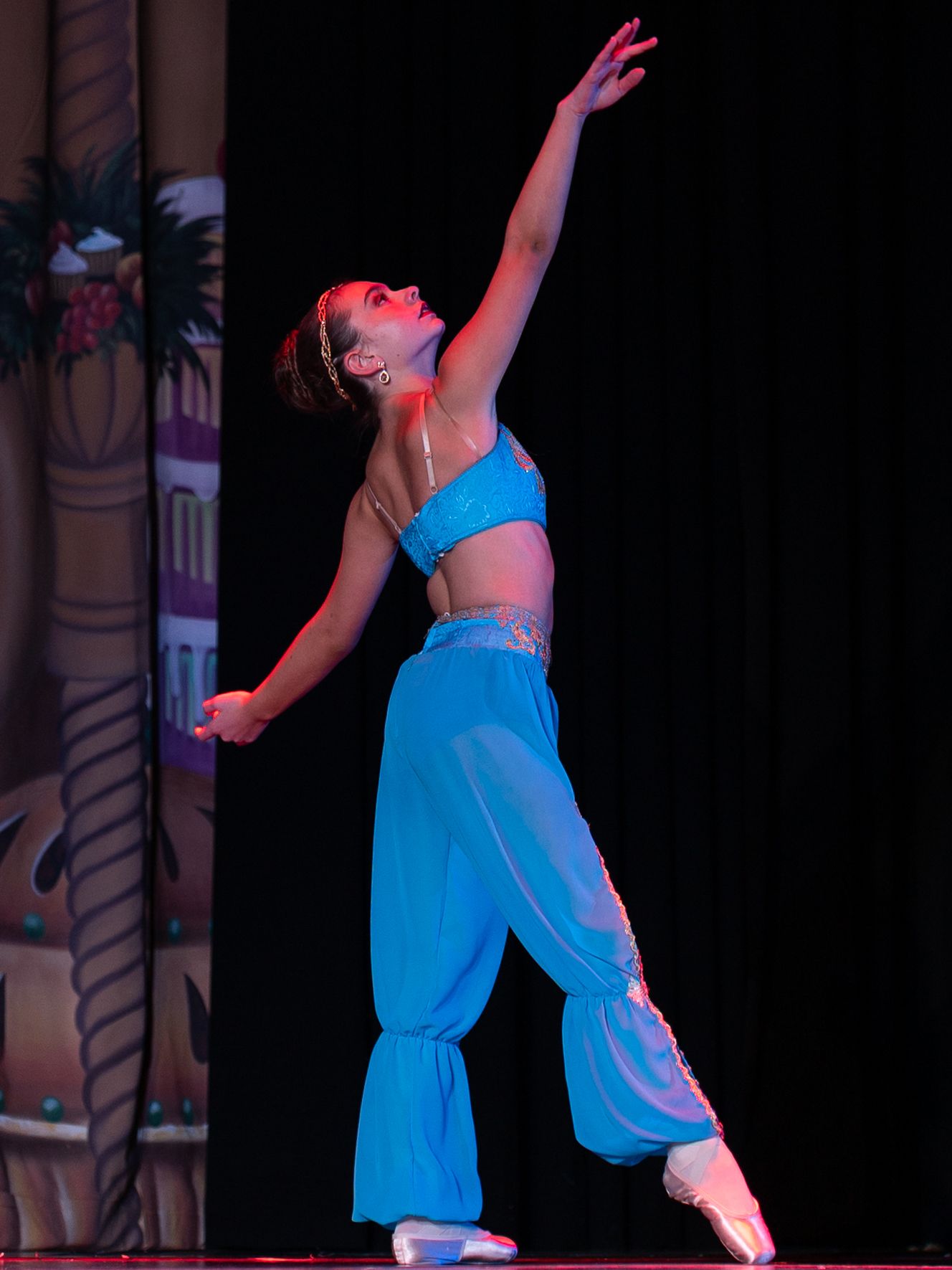 A young female ballet dancer performs on stage in a blue costume, elegantly reaching upward on pointe.