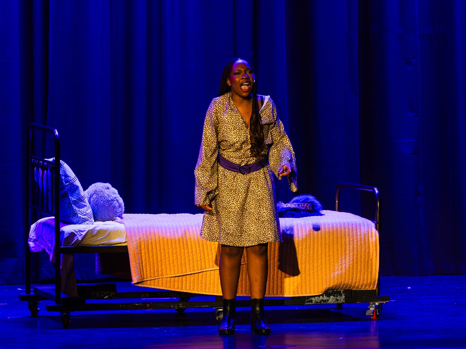 A woman in a floral dress performs a dramatic solo scene beside a bed on a blue-lit stage.
