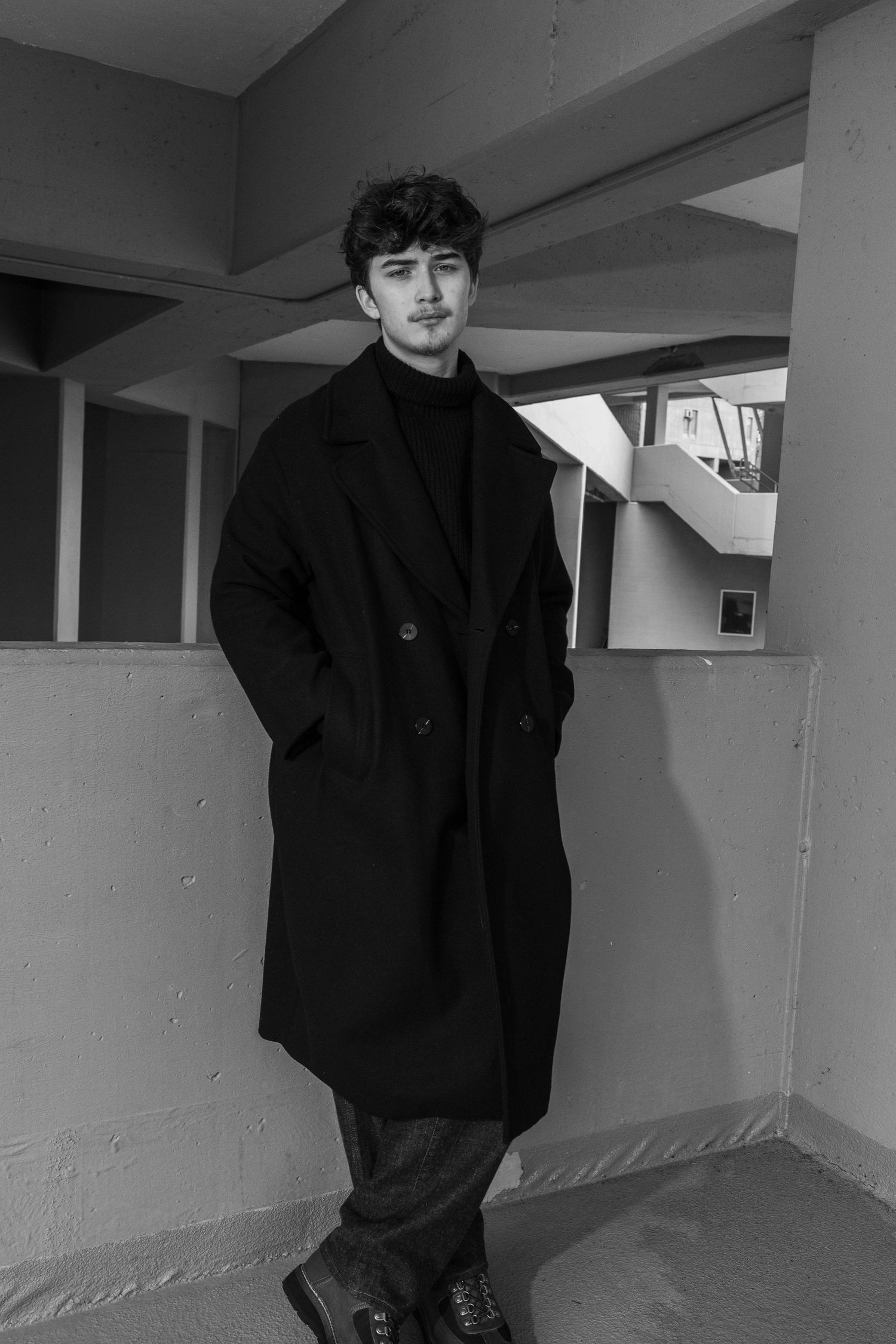 Black and white portrait of a young man with dark curly hair in a long dark overcoat and turtleneck, standing in a concrete building.