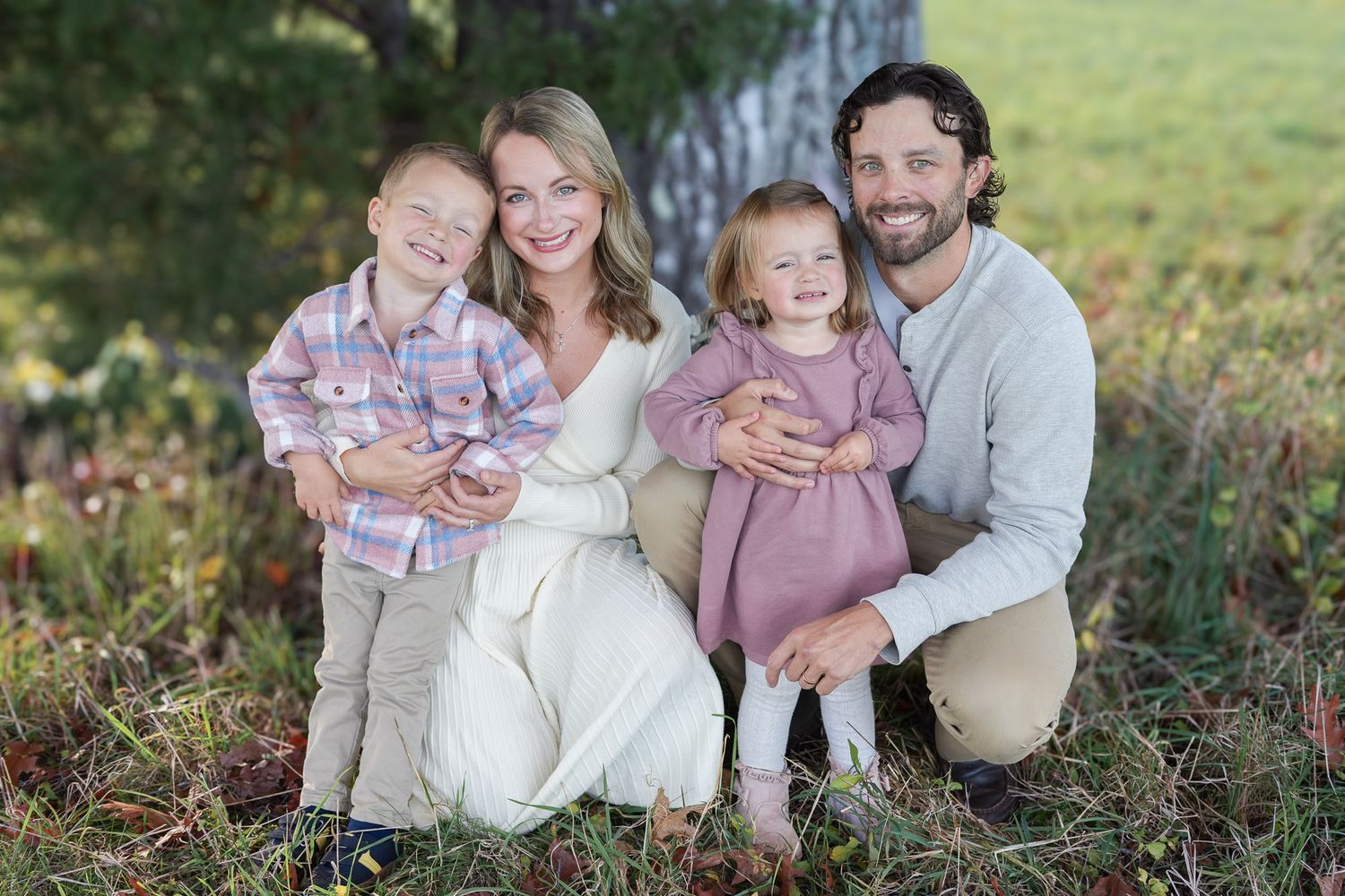 Smiling family of four posing together near a tree in an autumn outdoor setting.