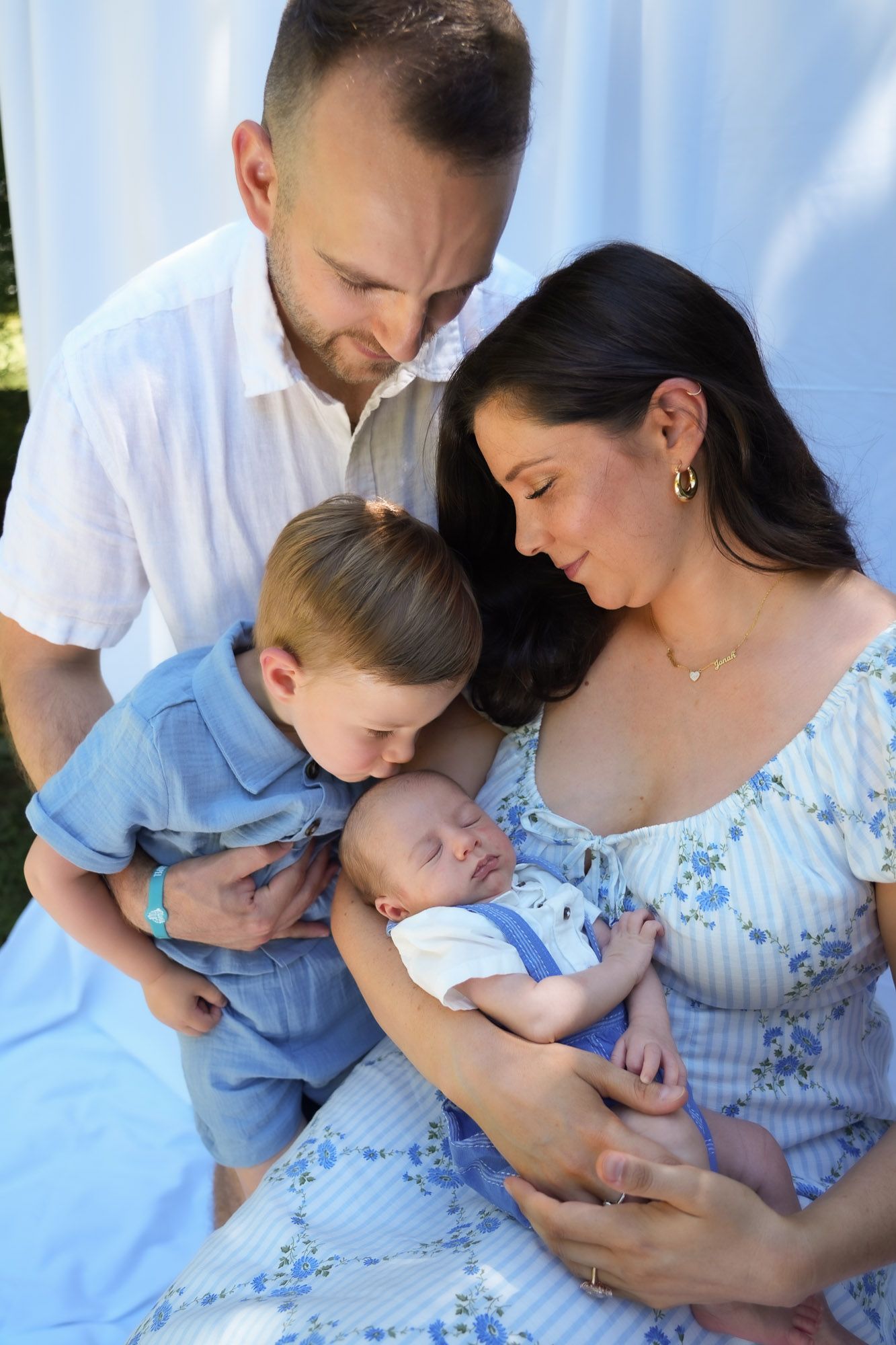 Family of four tenderly gathered around sleeping newborn baby in outdoor portrait.
