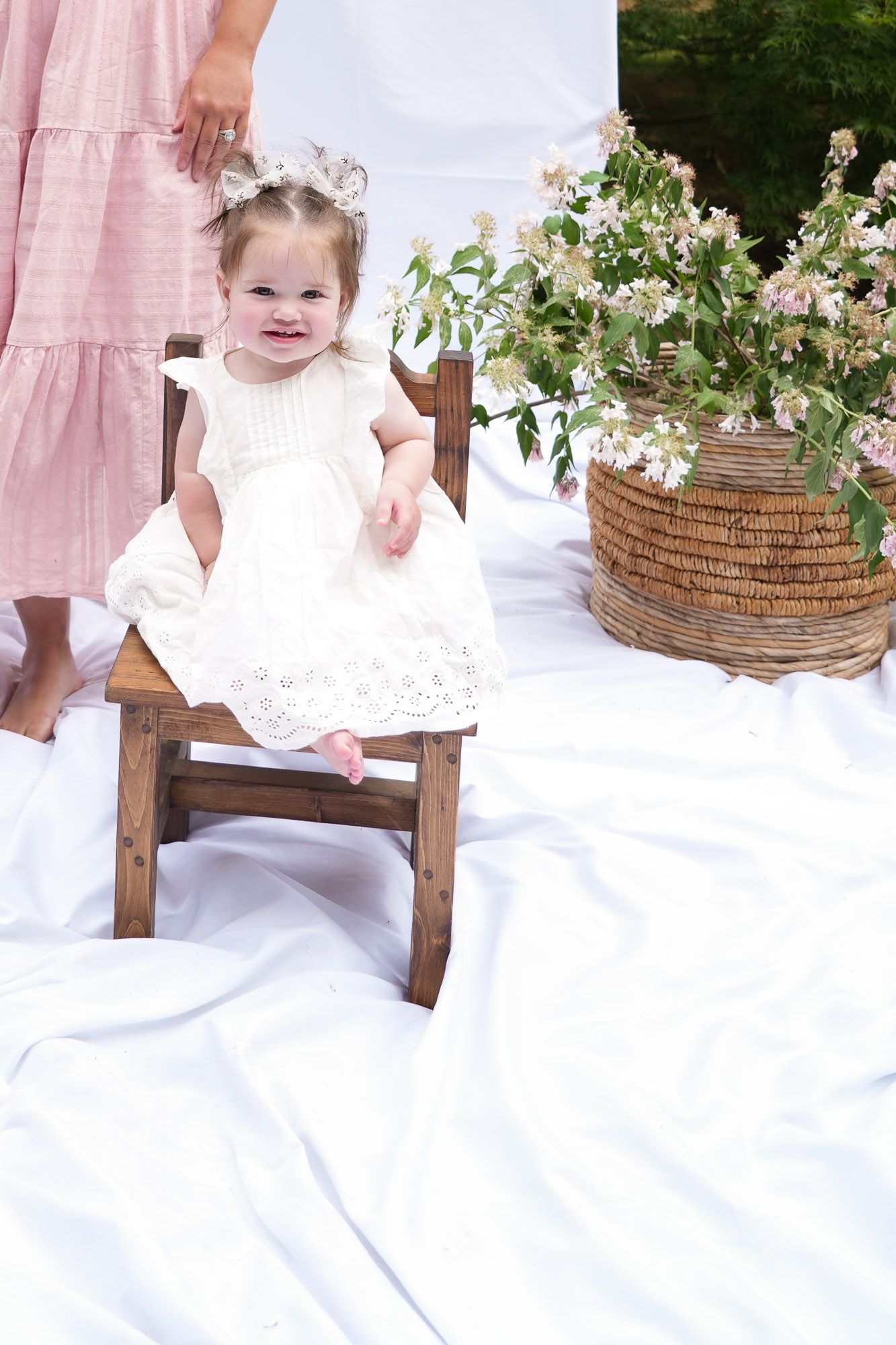 Adorable toddler girl in white lace dress sits on small wooden chair outdoors next to floral basket arrangement.