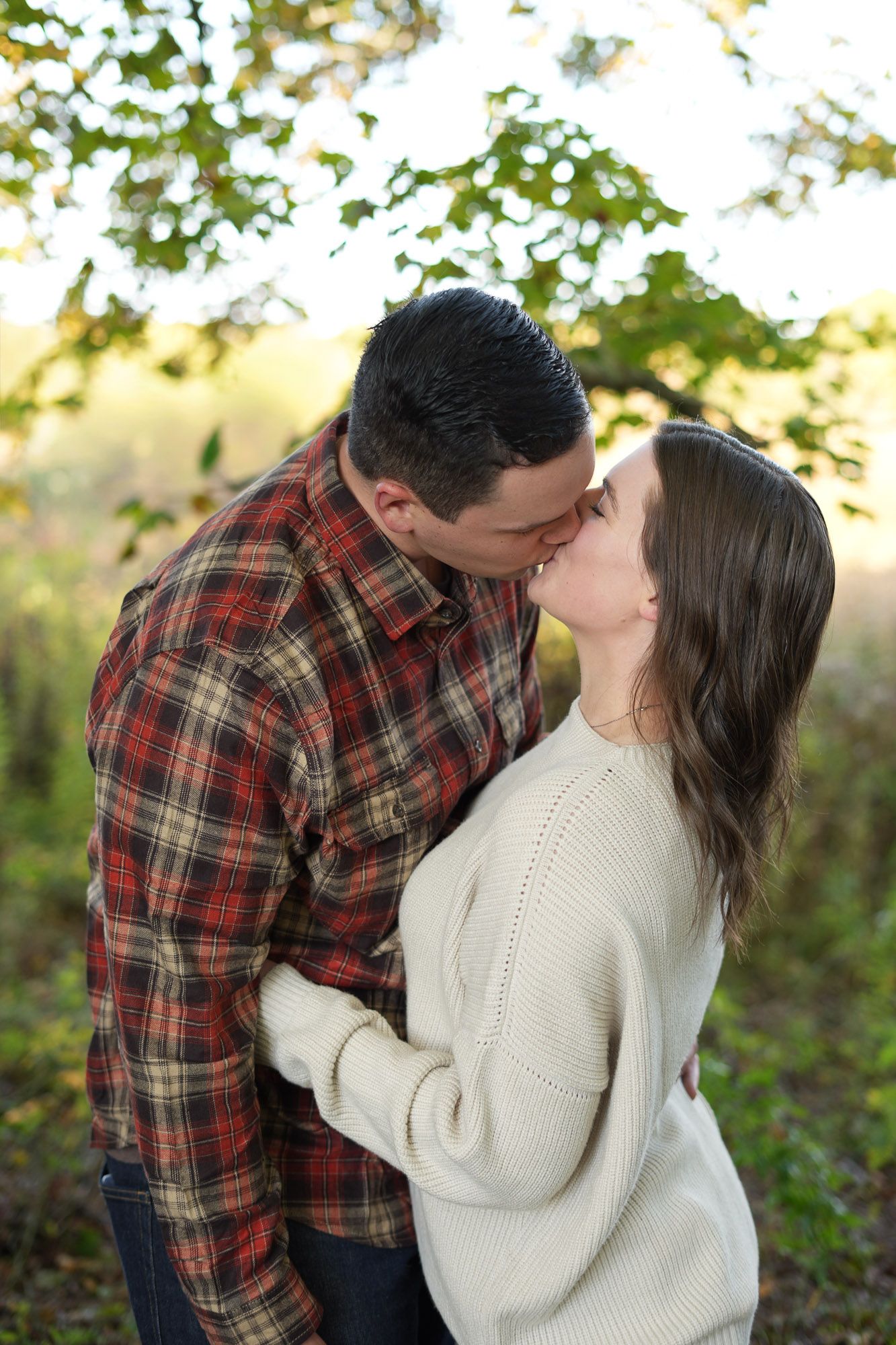 Couple kissing tenderly outdoors beneath a tree with autumn leaves, dressed in plaid and cream sweater.