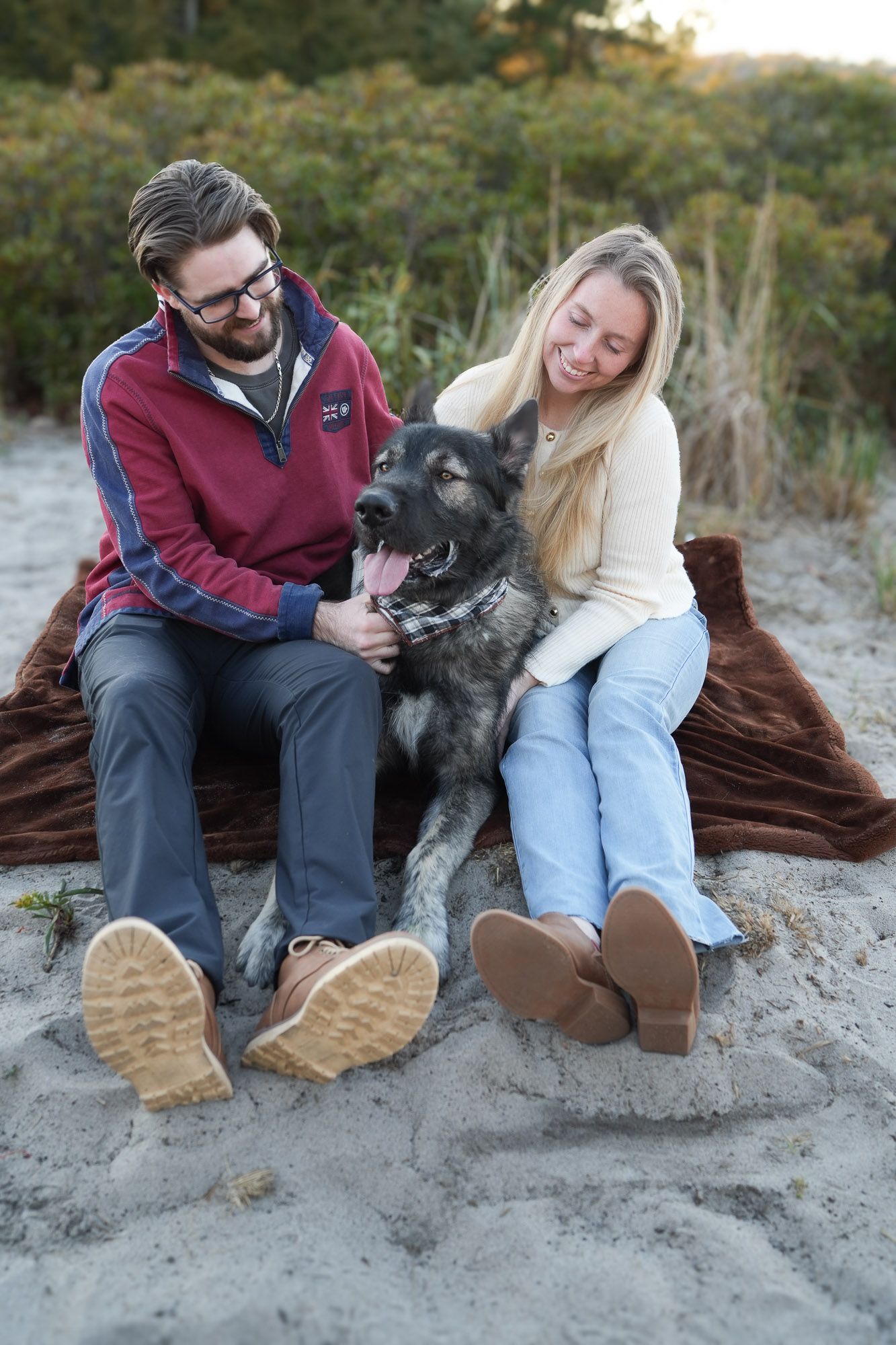Smiling couple with their panting German Shepherd dog sitting on a blanket on a sandy beach.