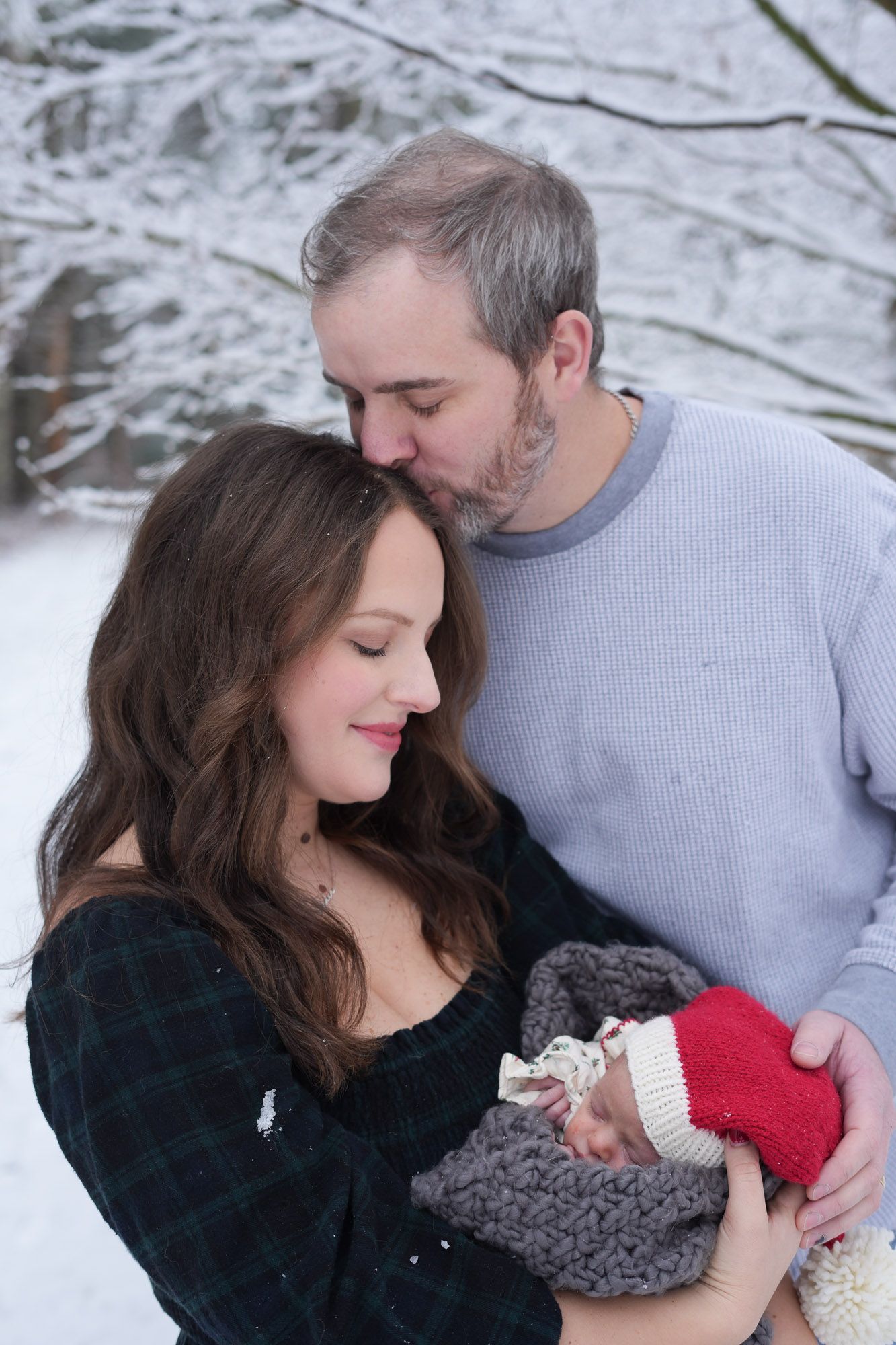 A loving couple shares a tender moment in a snowy forest, with the man kissing the womans head as she holds their newborn.