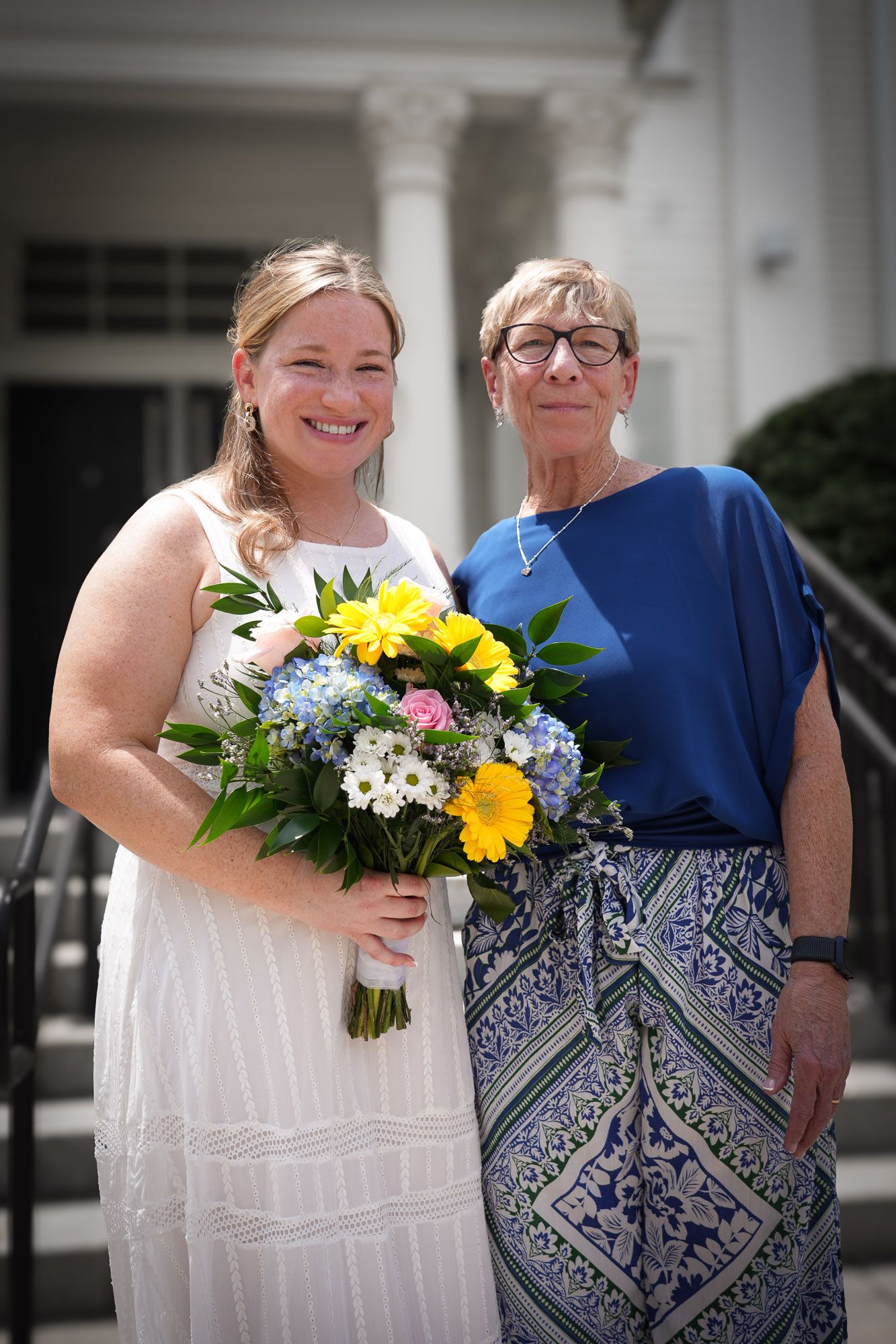 Bride in white dress holding colorful bouquet posing with older woman in blue outfit.