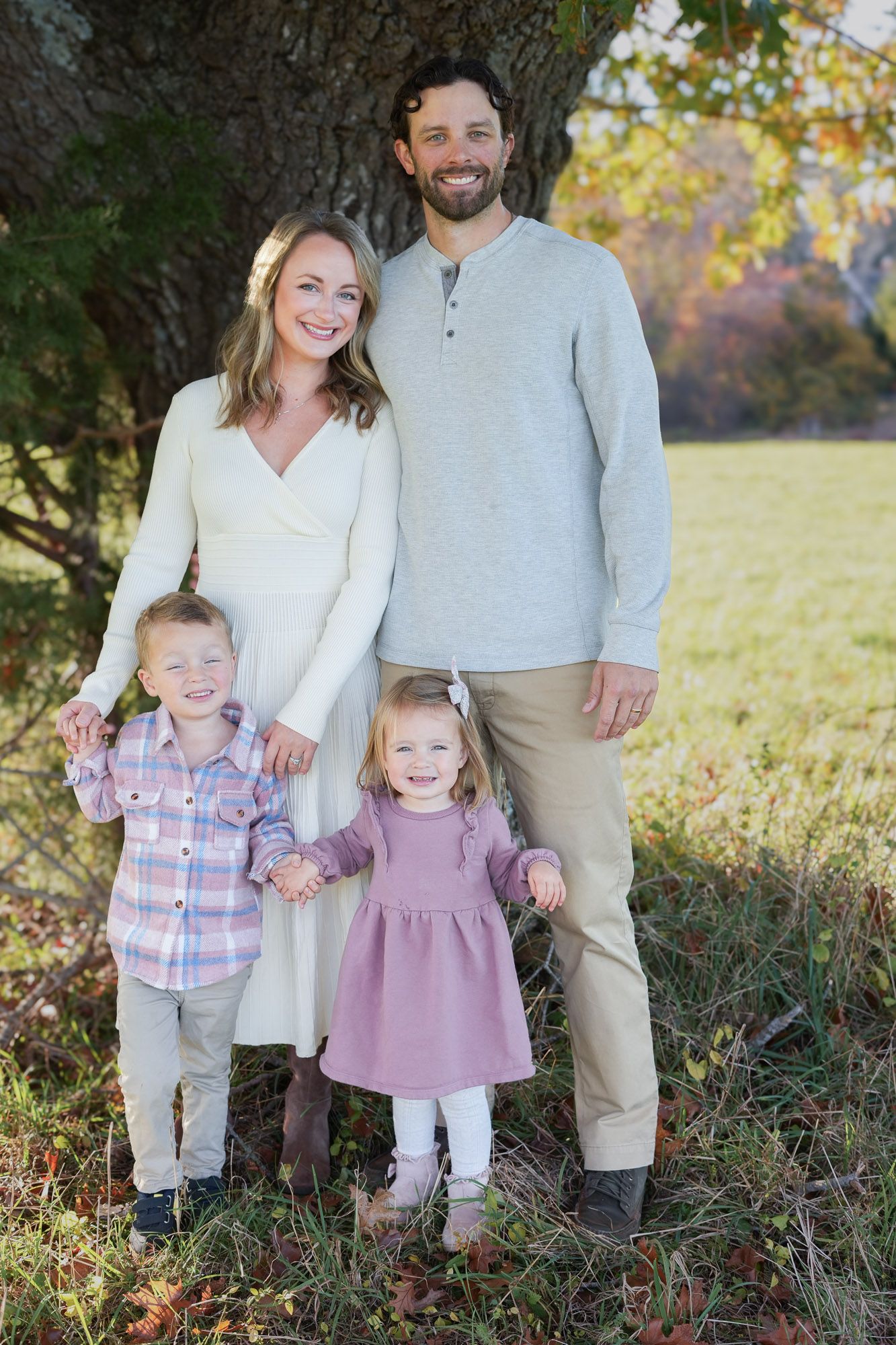 Happy family of four posing under a large tree in a park during autumn, smiling together.